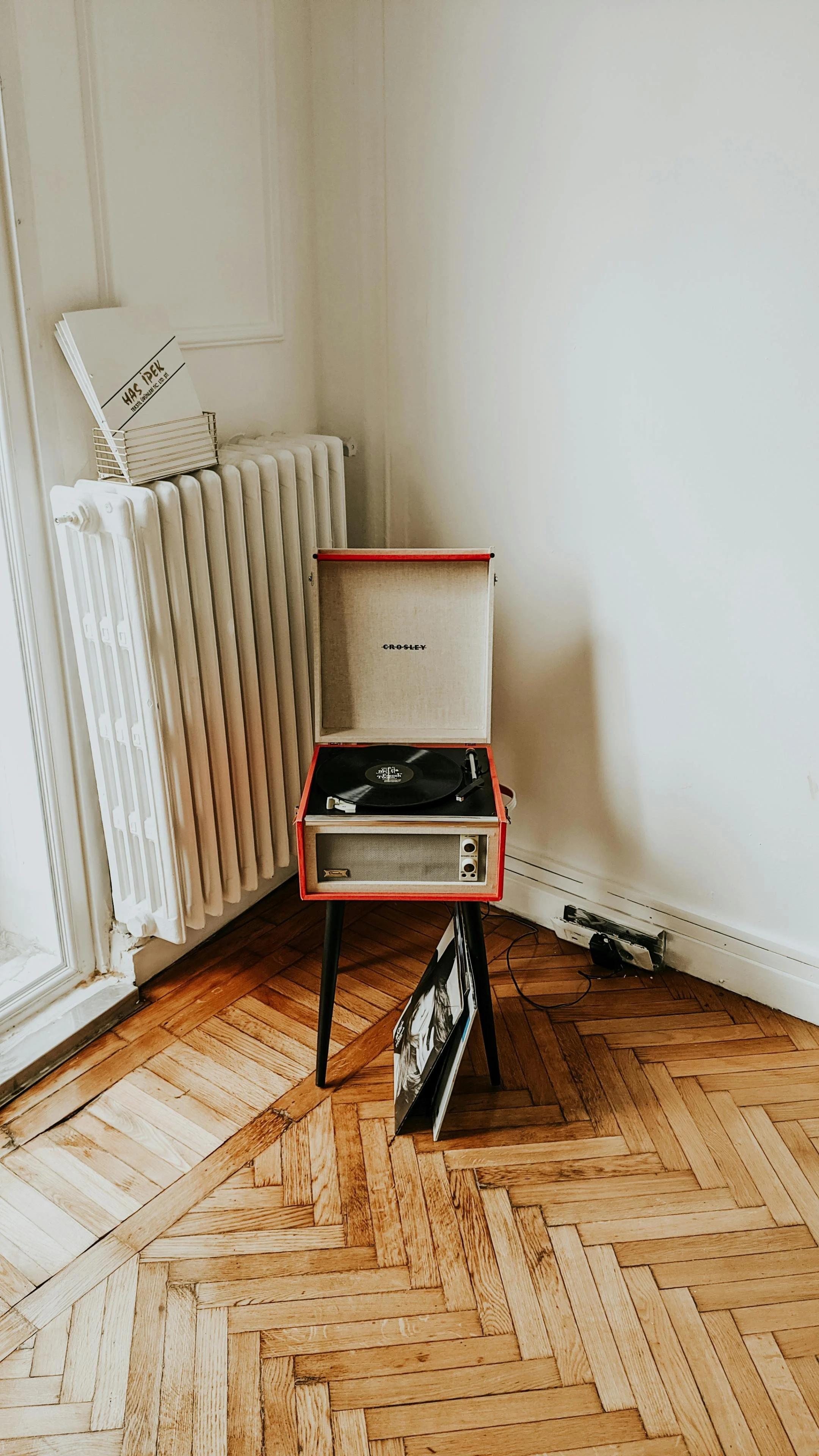 A vintage Crosley record player on a small black table, with a record playing. Next to it is a framed photo leaning against the table. In the background, there is a white radiator and a window ledge with a wire basket containing papers and a sign tha
