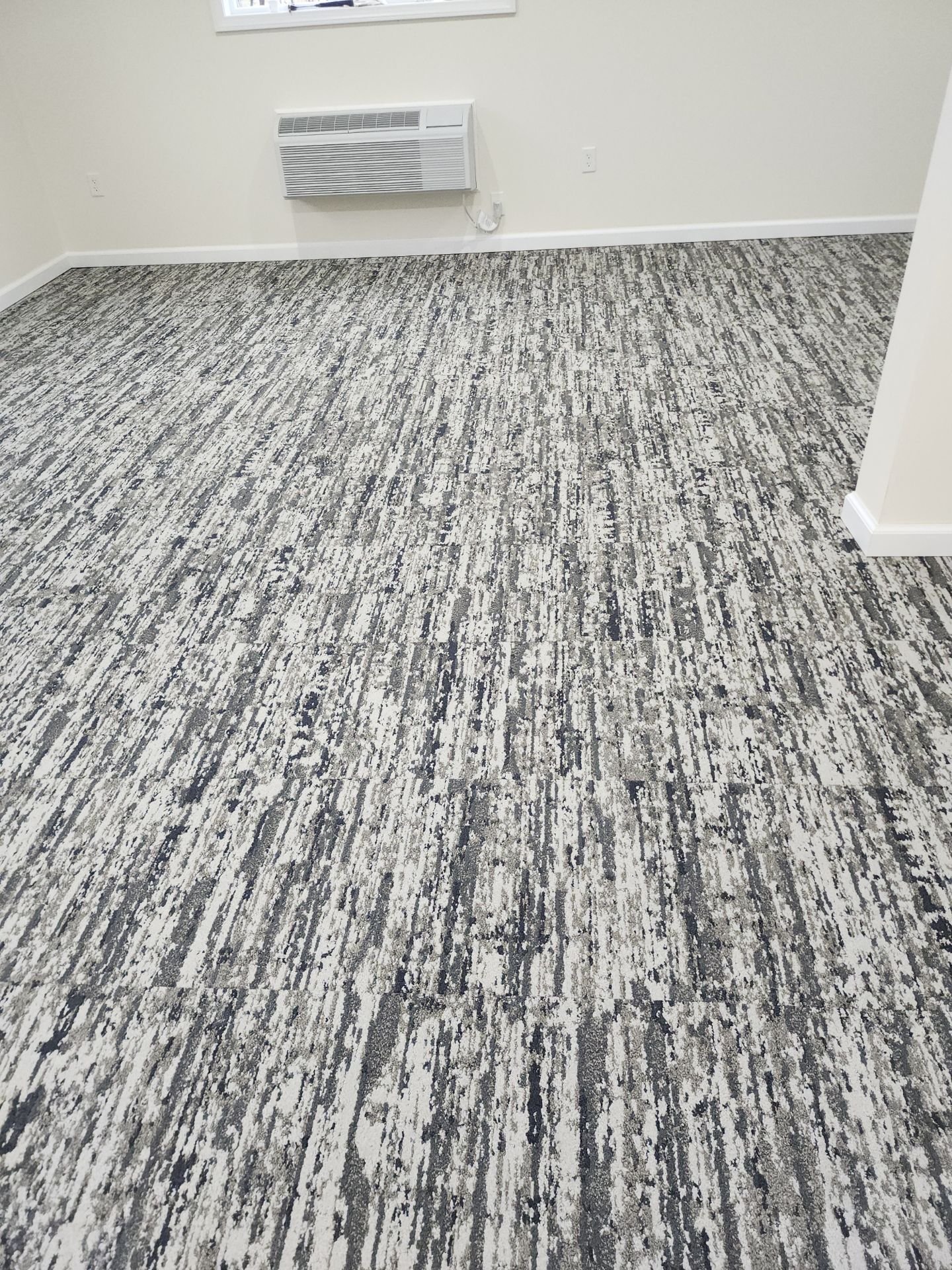 Empty room with patterned gray, black, and white carpet, beige walls, white baseboards, and a wall-mounted HVAC unit.