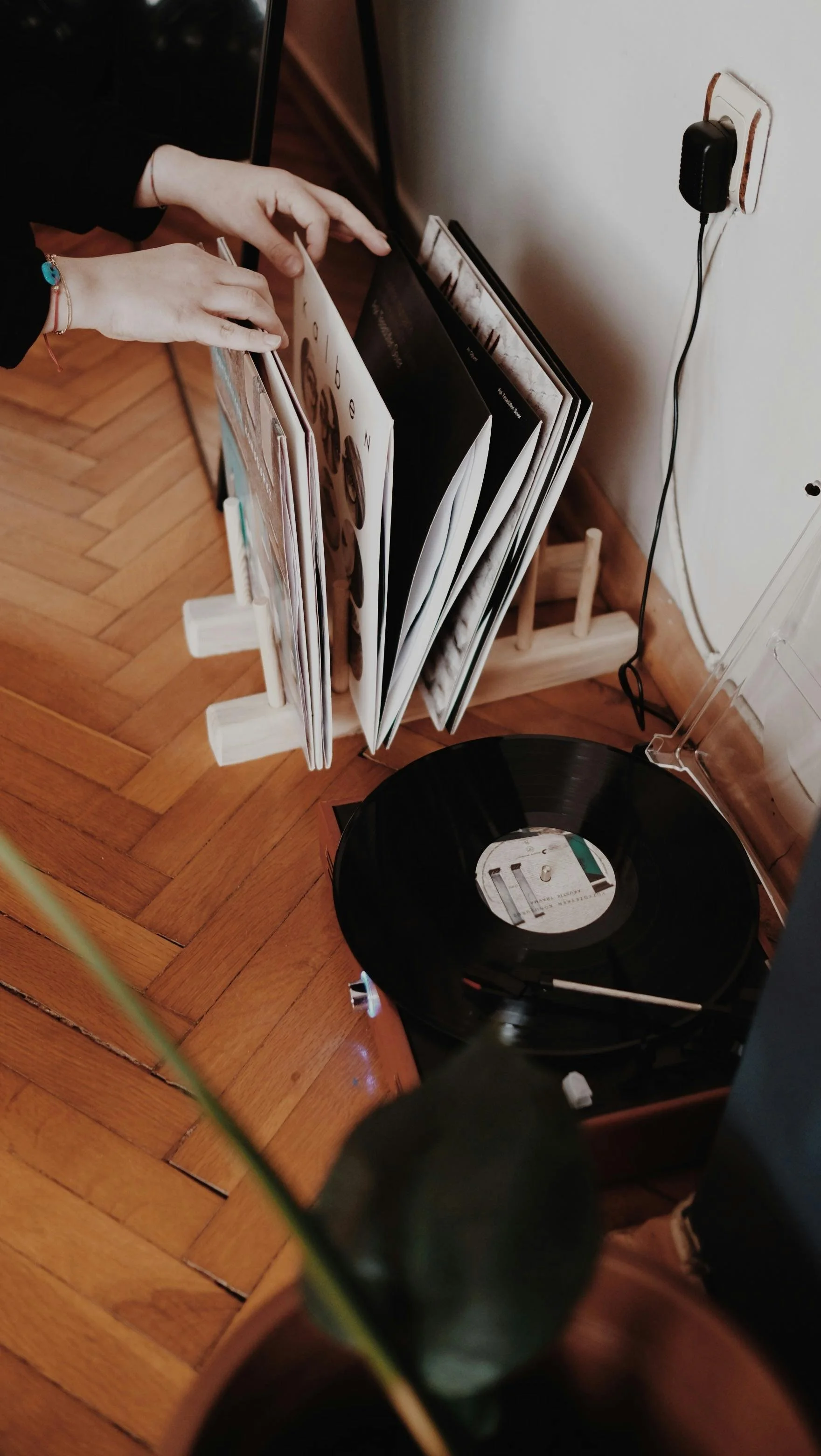 A person with a bracelet on their wrist is removing or organizing magazines or vinyl records from a wooden rack beside a turntable with a black vinyl record, placed on a wooden surface.