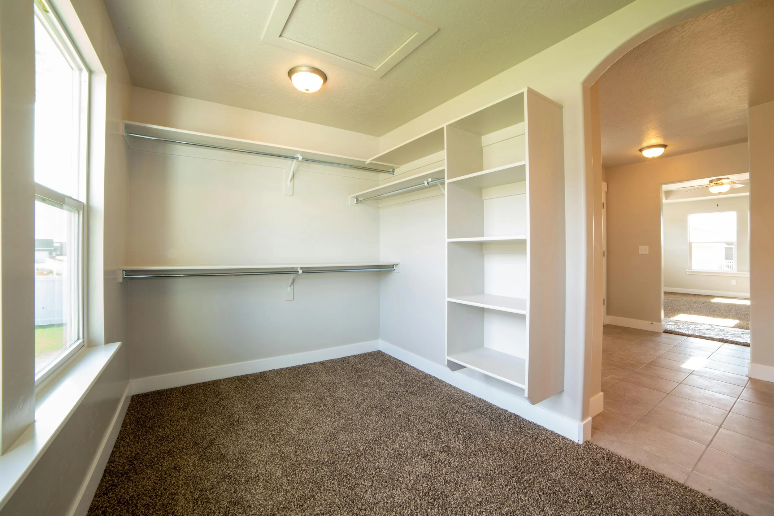 Empty walk-in closet with white shelving and hanging rods, beige carpeting, two large windows, and view into a room with tile flooring and a window.