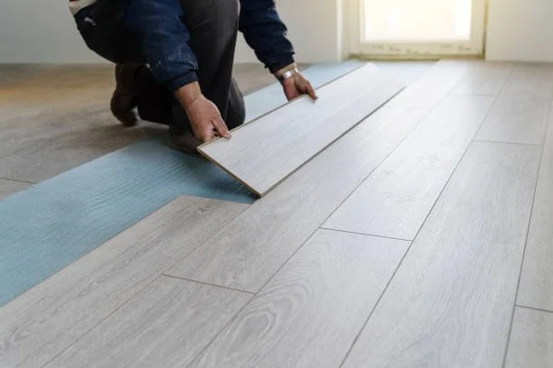 Person installing light-colored wood or laminate flooring in a room with a window