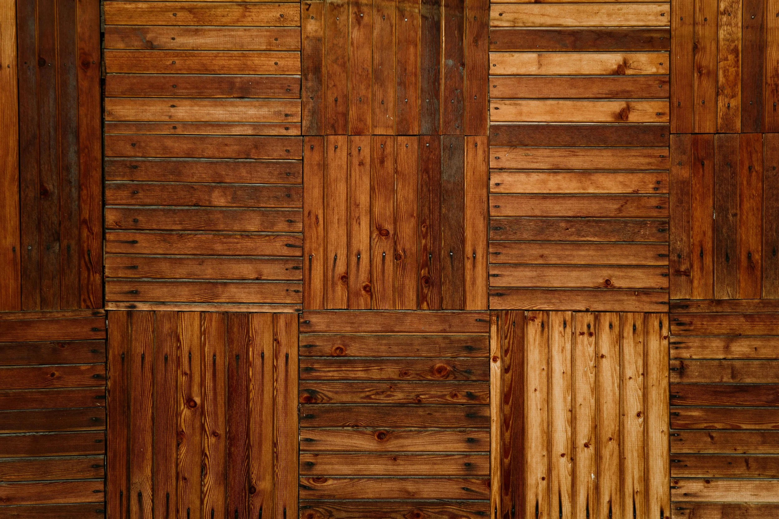 Close-up of a wooden wall made up of square panels of reddish-brown wood with visible grain patterns and small nails.