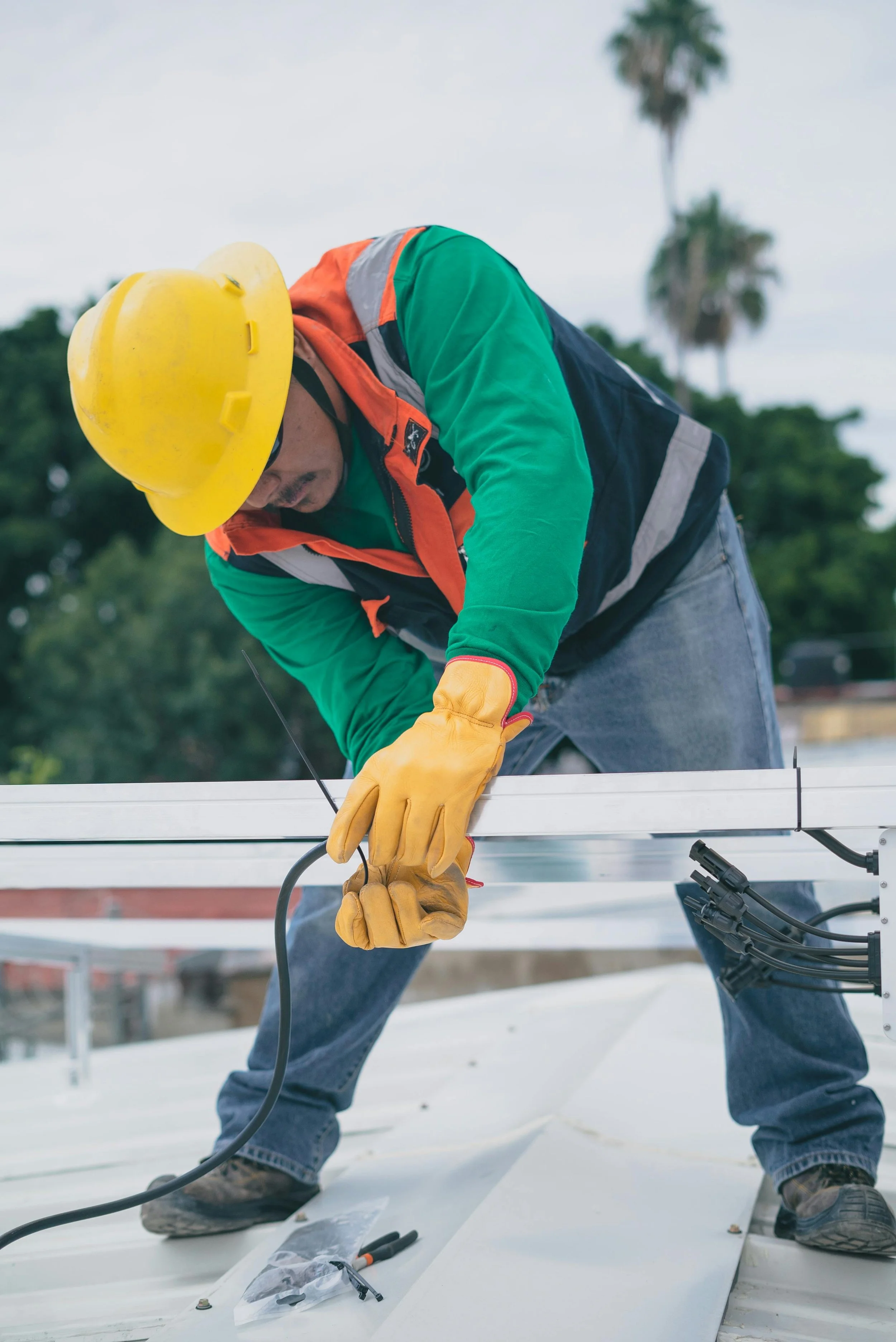 A construction worker wearing a yellow hard hat, yellow gloves, green shirt, and high visibility vest is installing or repairing a white metal structure on a roof. There are palm trees in the background.
