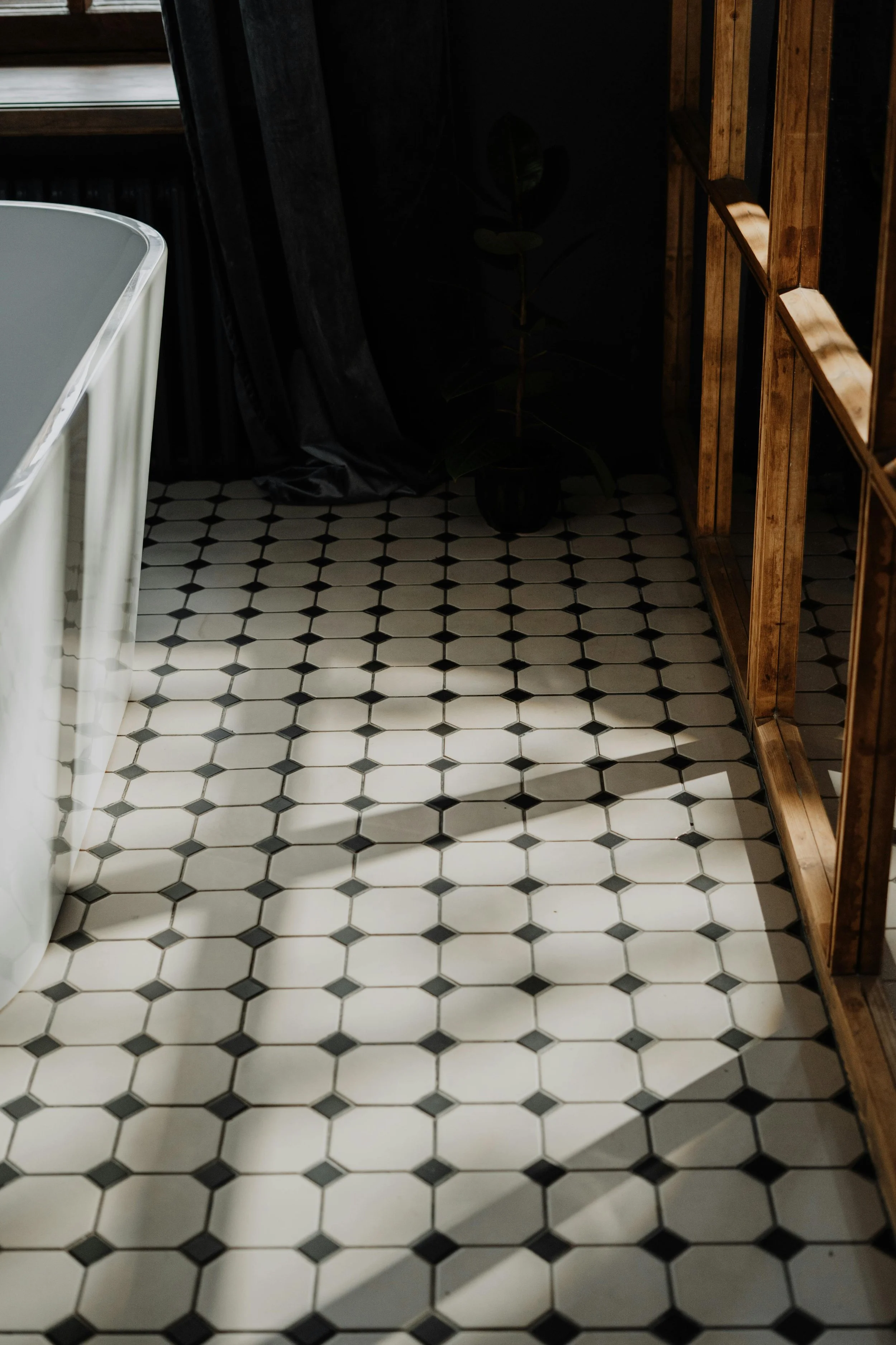 Close-up of a tiled black and white floor with shadows cast across it, adjacent to a wooden shelving unit and a potted plant in a black container in a dimly lit room.