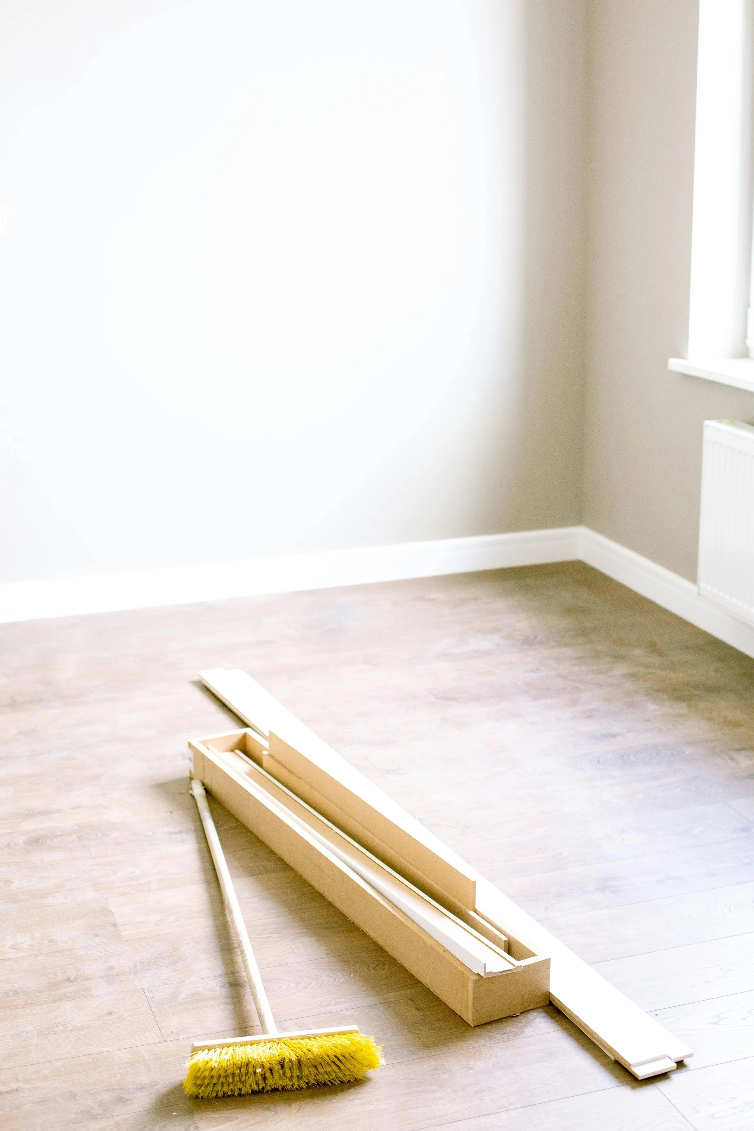 An empty room with light-colored walls and wooden flooring, featuring a broom and assembled furniture parts on the floor near a window.