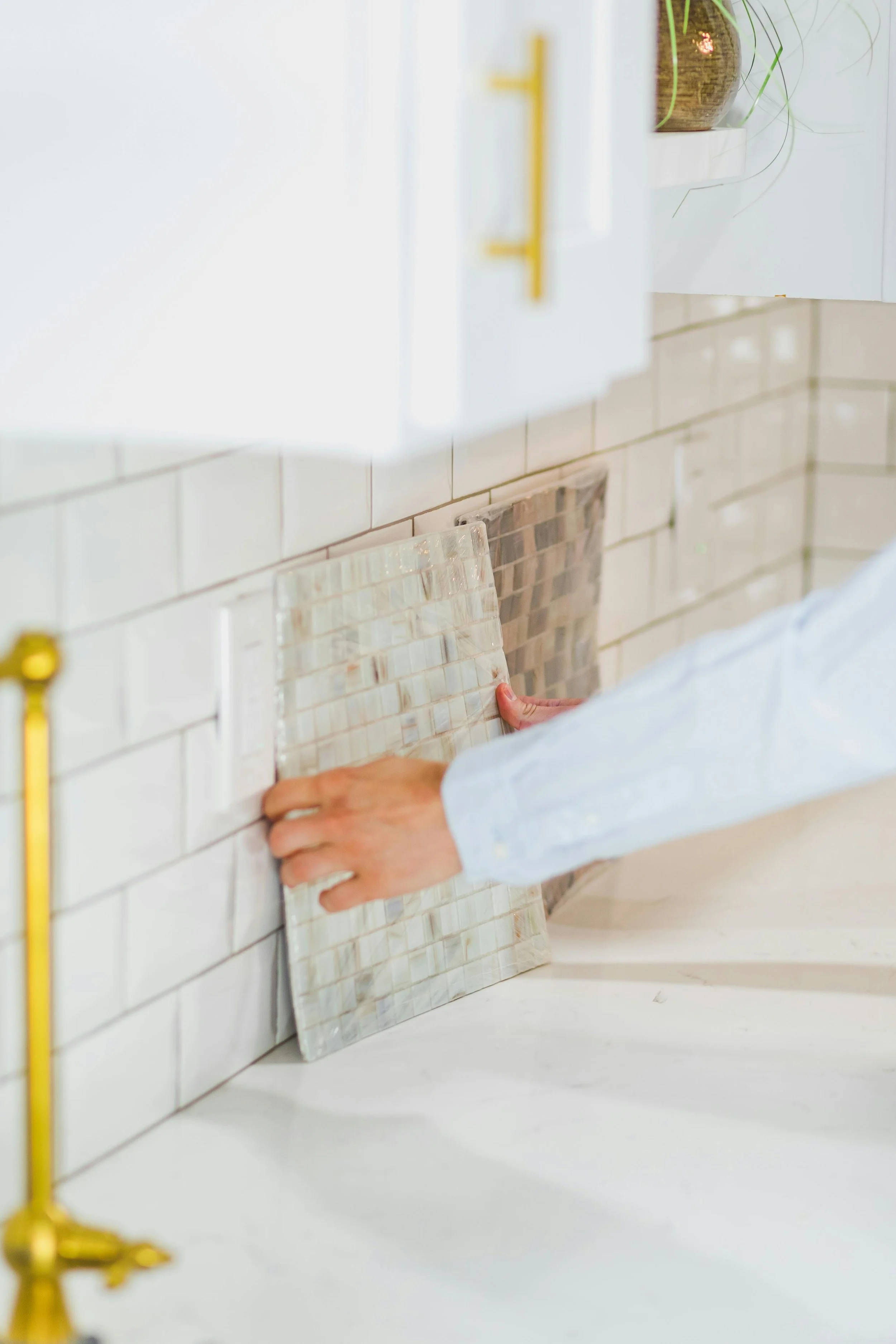 Person installing a tile backsplash in a kitchen with white cabinets, beige brick wall, and decorative pots.