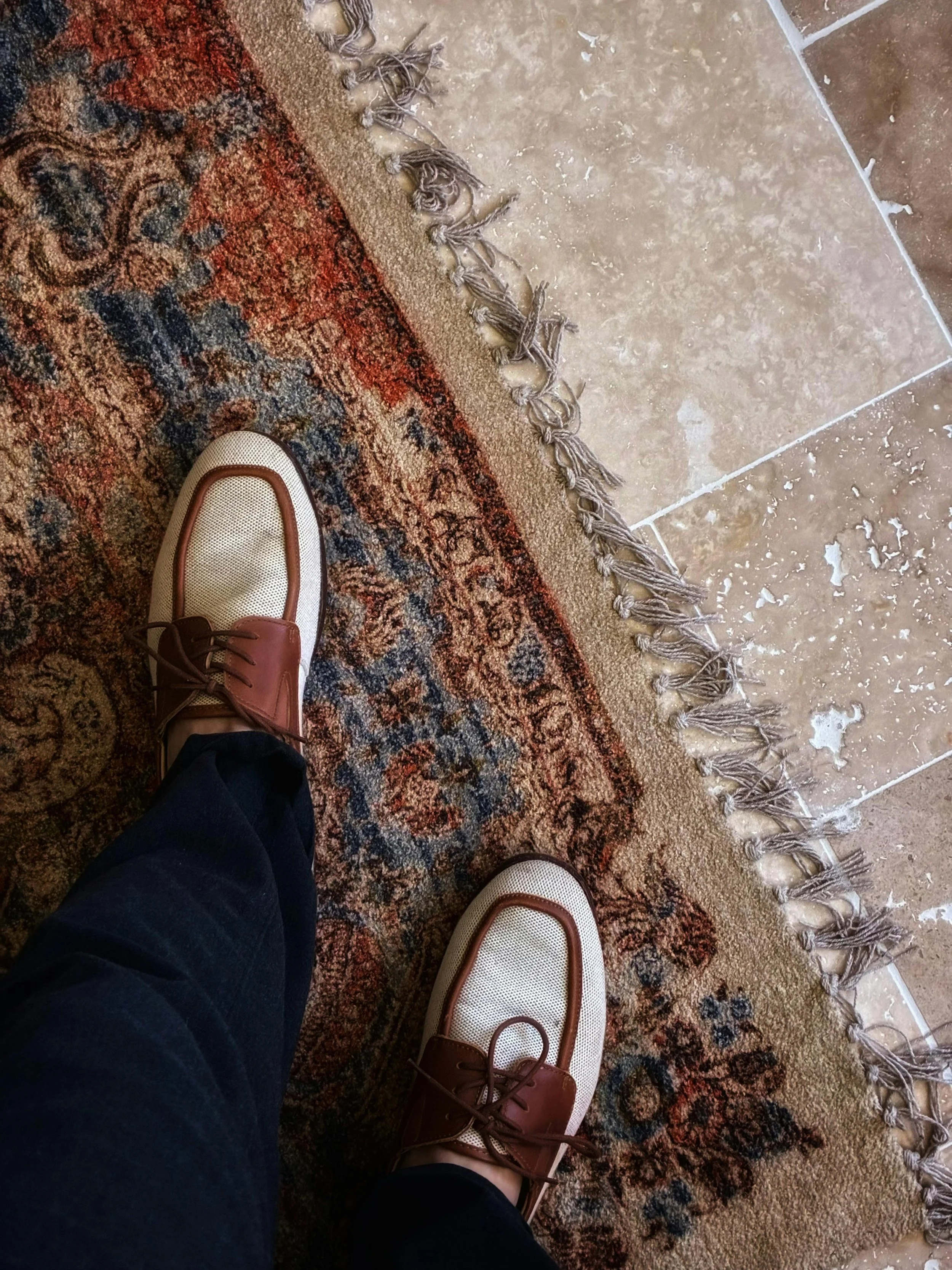 View of a person's feet wearing beige shoes with brown laces, standing on a patterned rug near a tiled floor with a decorative fringe.
