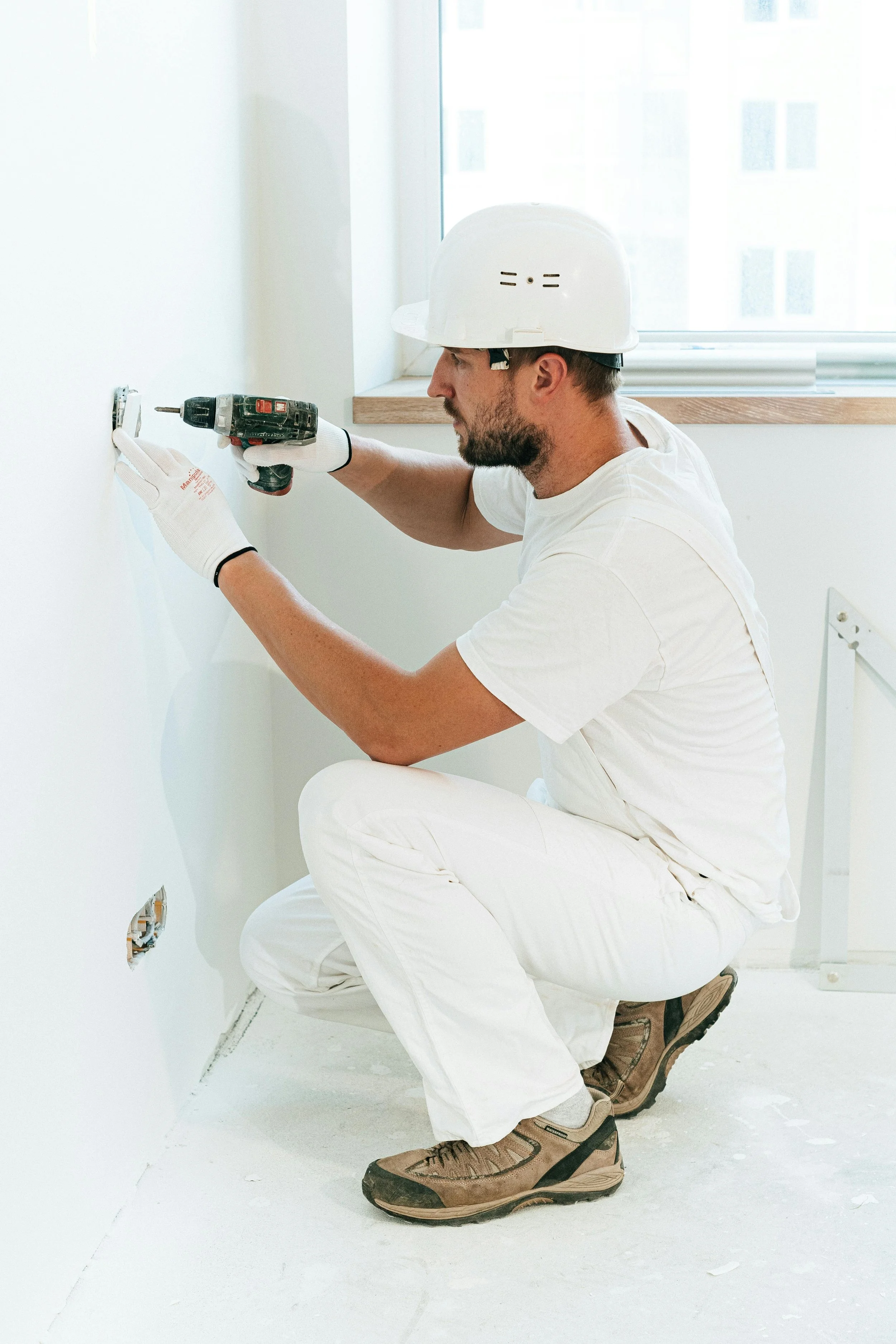 Man wearing a white hard hat and gloves installing an electrical outlet in a white wall using a drill.