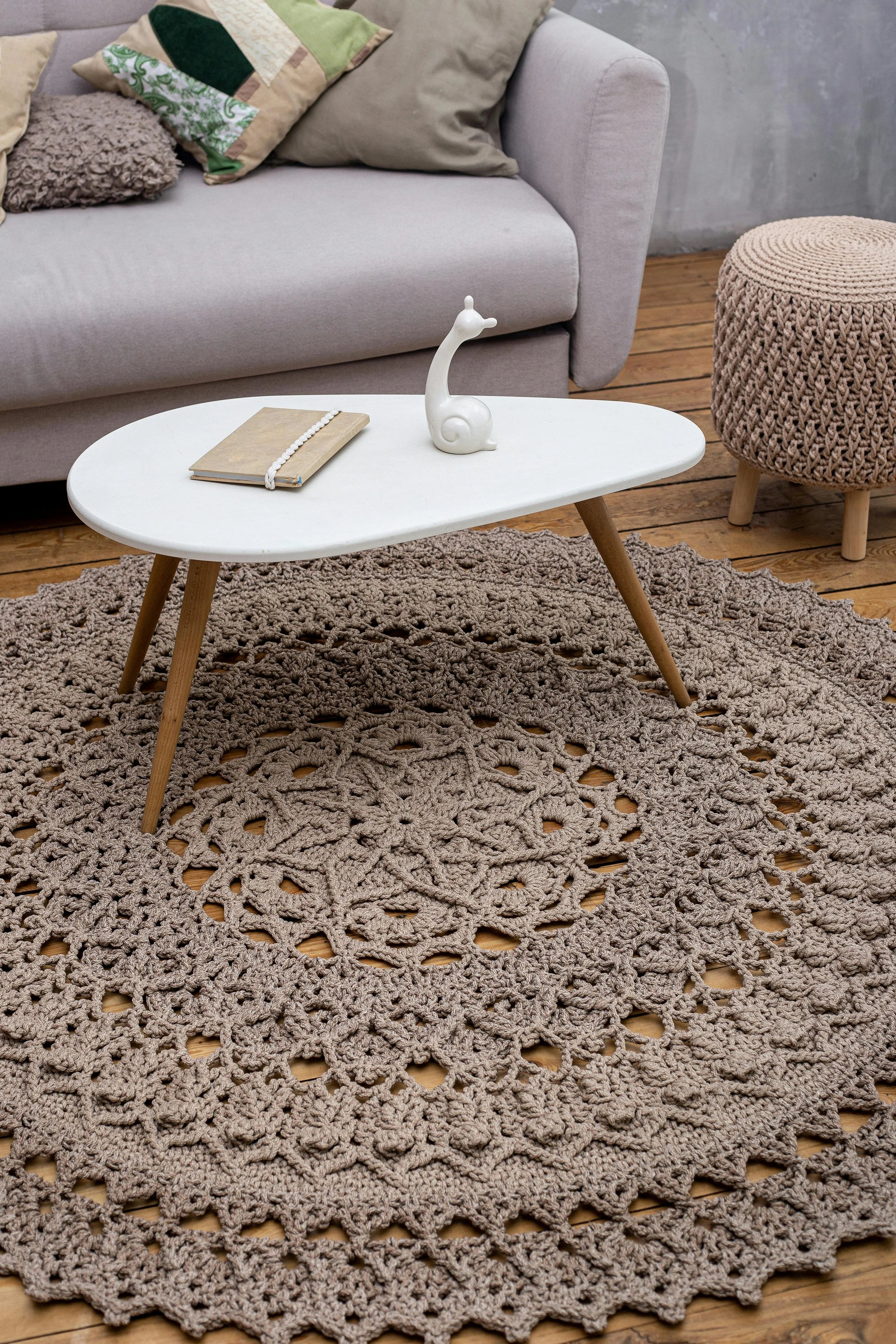 Living room with a beige sofa, multicolored pillows, a round knitted ottoman, a white coffee table with a decorative ceramic giraffe and a book, a crocheted rug on a wooden floor, and a gray textured wall.