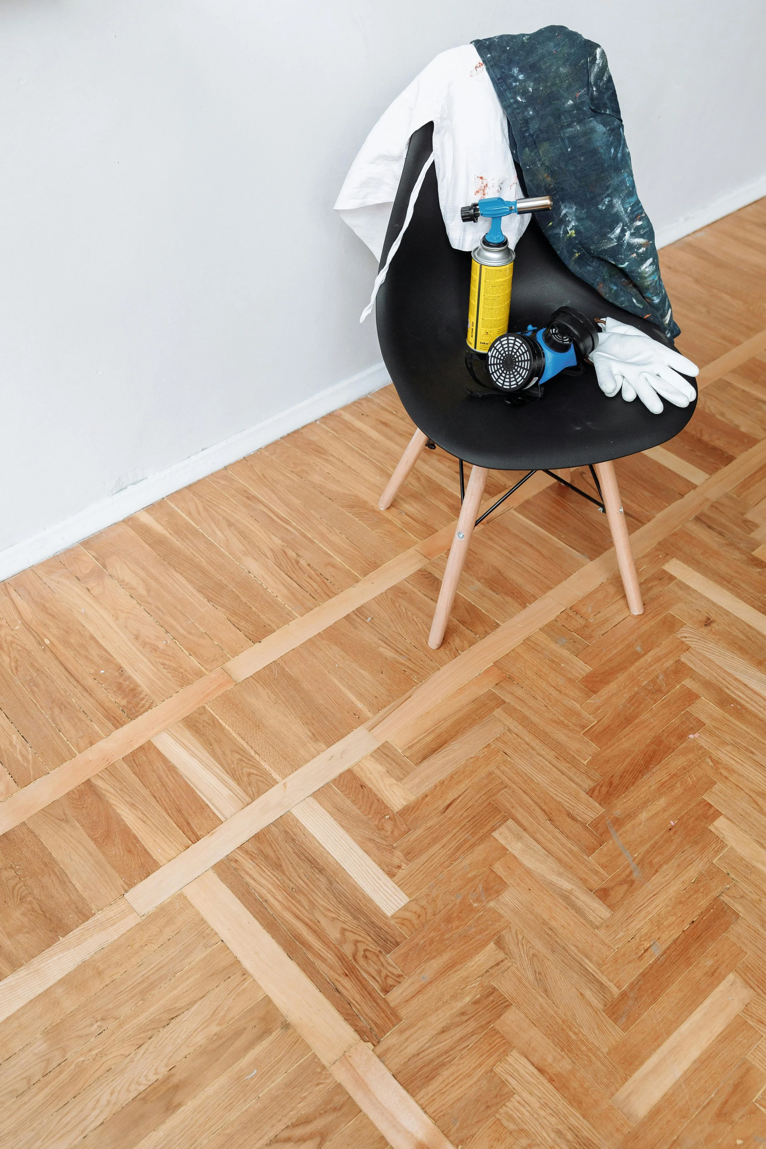 A black chair with wooden legs holding a paint spray can, a respirator, white gloves, and a cloth draped over the back, on a hardwood floor with in-progress floor refinishing.