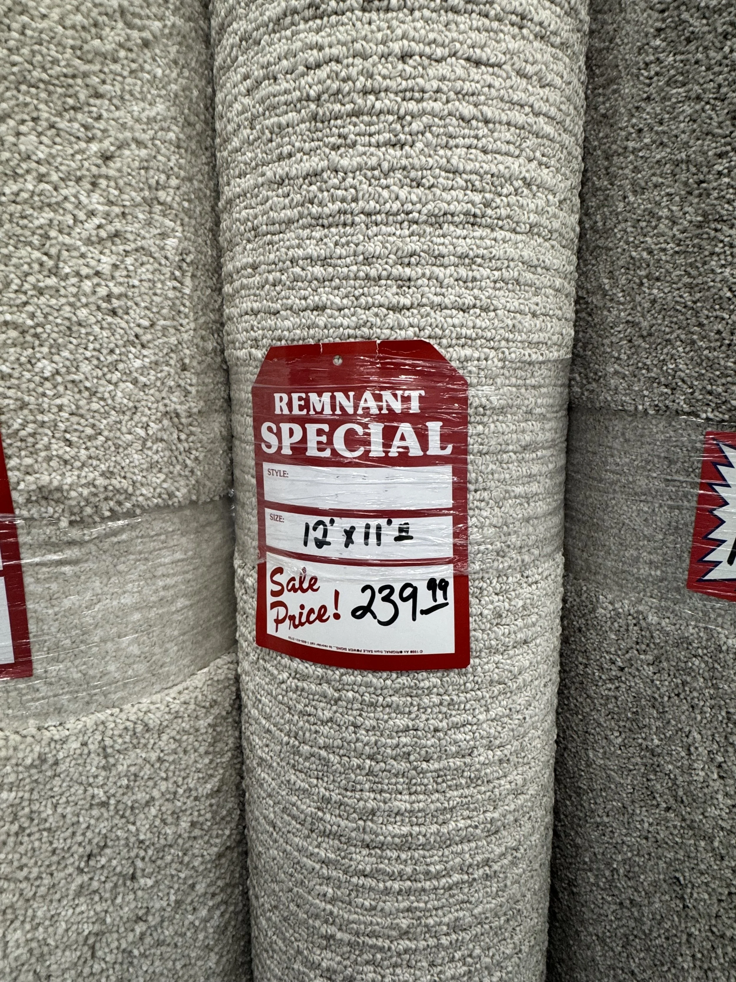 Rolled beige and gray carpets in a store with a red and white sale tag indicating size 12' x 10' and a price of $239.99.