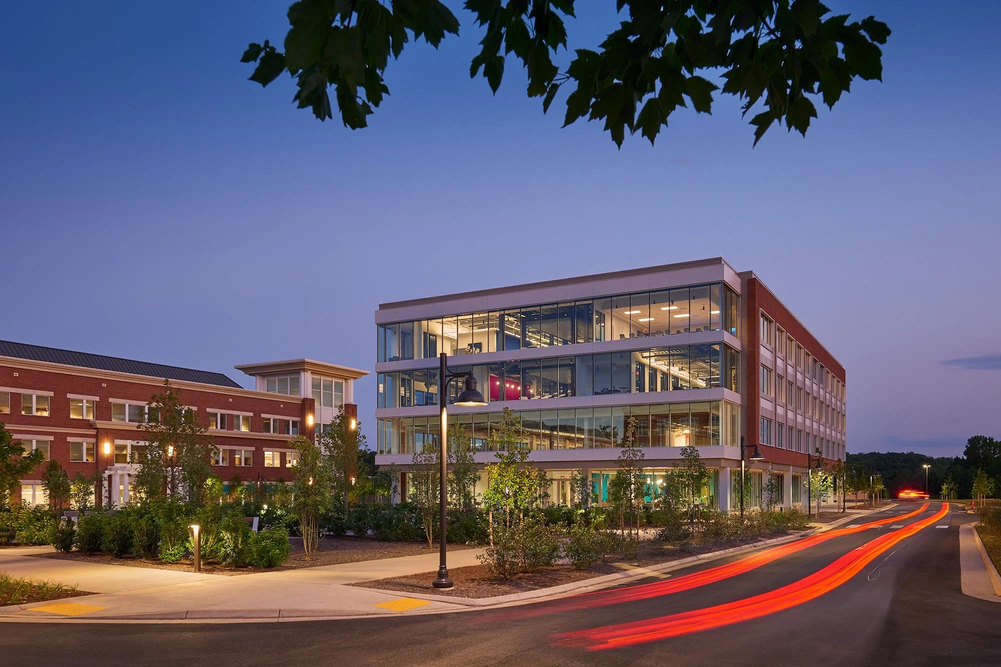 Modern multi-story office building with glass windows at dusk, surrounded by landscaped greenery and a curved road with light trails from passing cars.