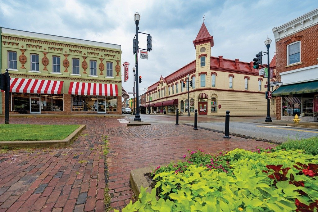 Empty historic downtown street with brick sidewalks, small shops, and colorful buildings, including a corner building with a tower, under a cloudy sky.