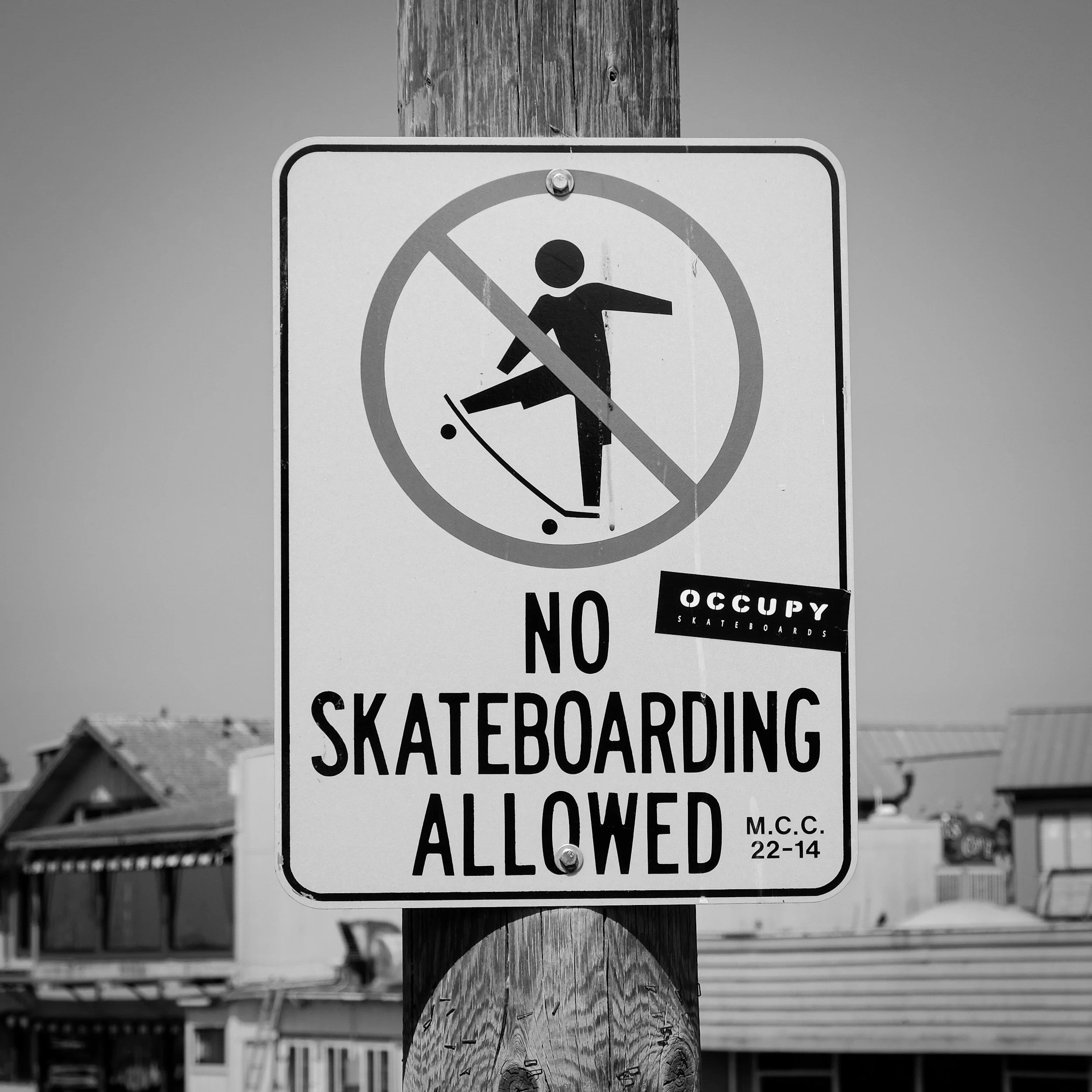 A black-and-white sign on a wooden pole indicating that skateboarding is not allowed, with a silhouette of a person skateboarding crossed out, and a sticker that says 'OCCUPY SKATEBOARDS'.