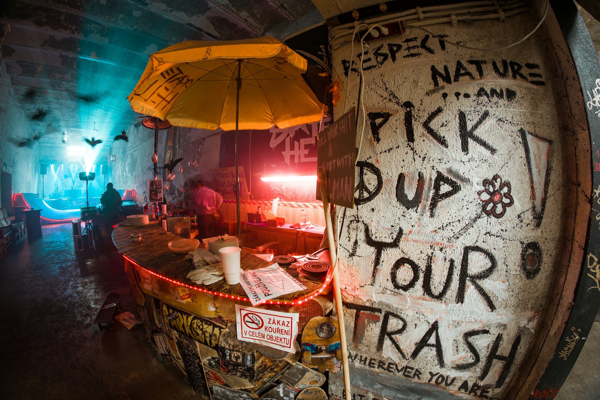 An indoor graffiti-covered space with a bar counter, a yellow umbrella, and a neon sign. Graffiti on the wall includes phrases like 'Respect Nature' and 'Pick Up!' Decorative bats hang from the ceiling. People are seen in the background, and there is a neon laser beam.