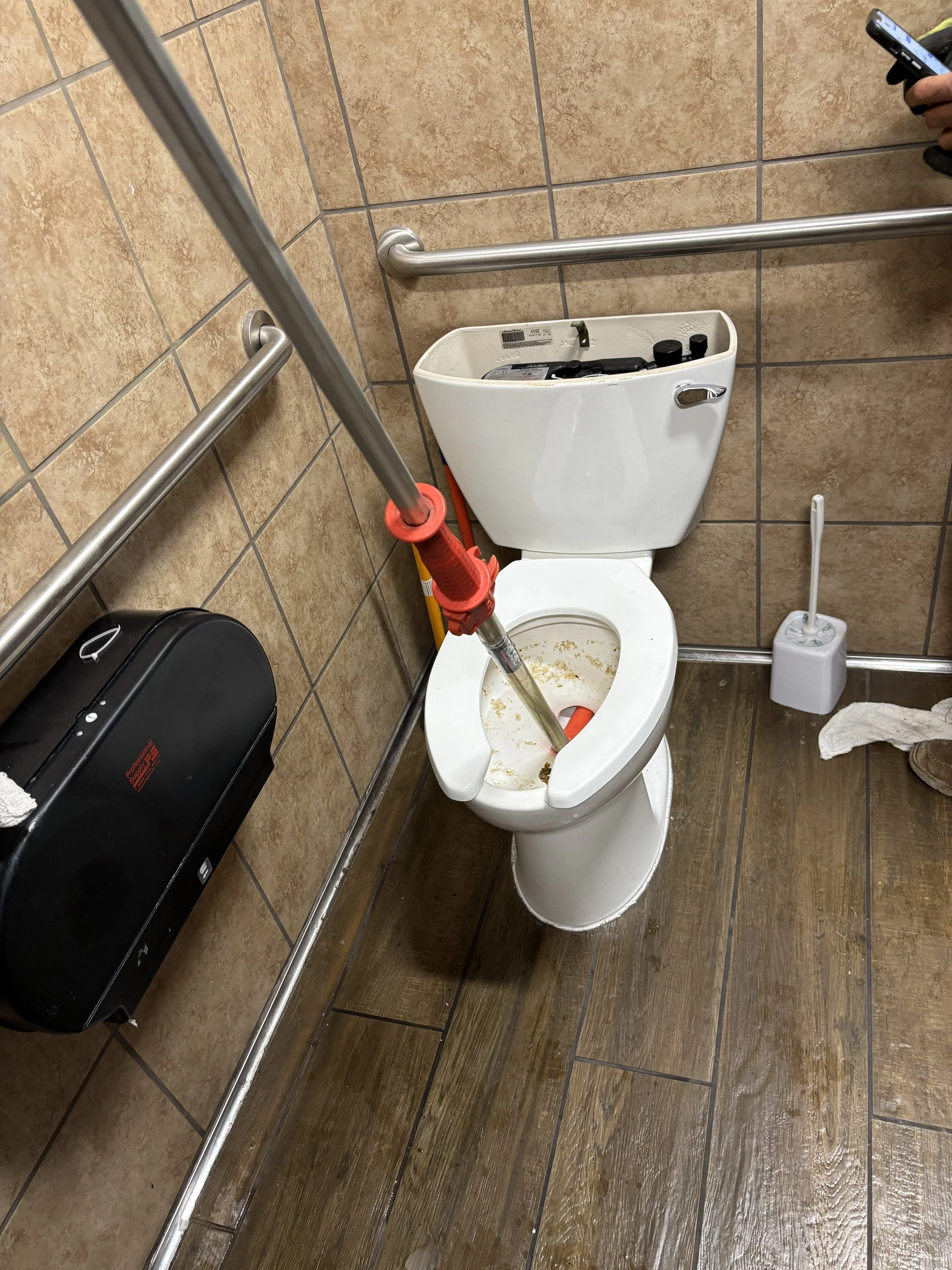 A bathroom stall with a toilet being cleaned with a toilet Auger. The toilet has some dirty water inside and cleaning supplies on the tank. Toilet paper holder, toilet brush, and wooden flooring are visible.