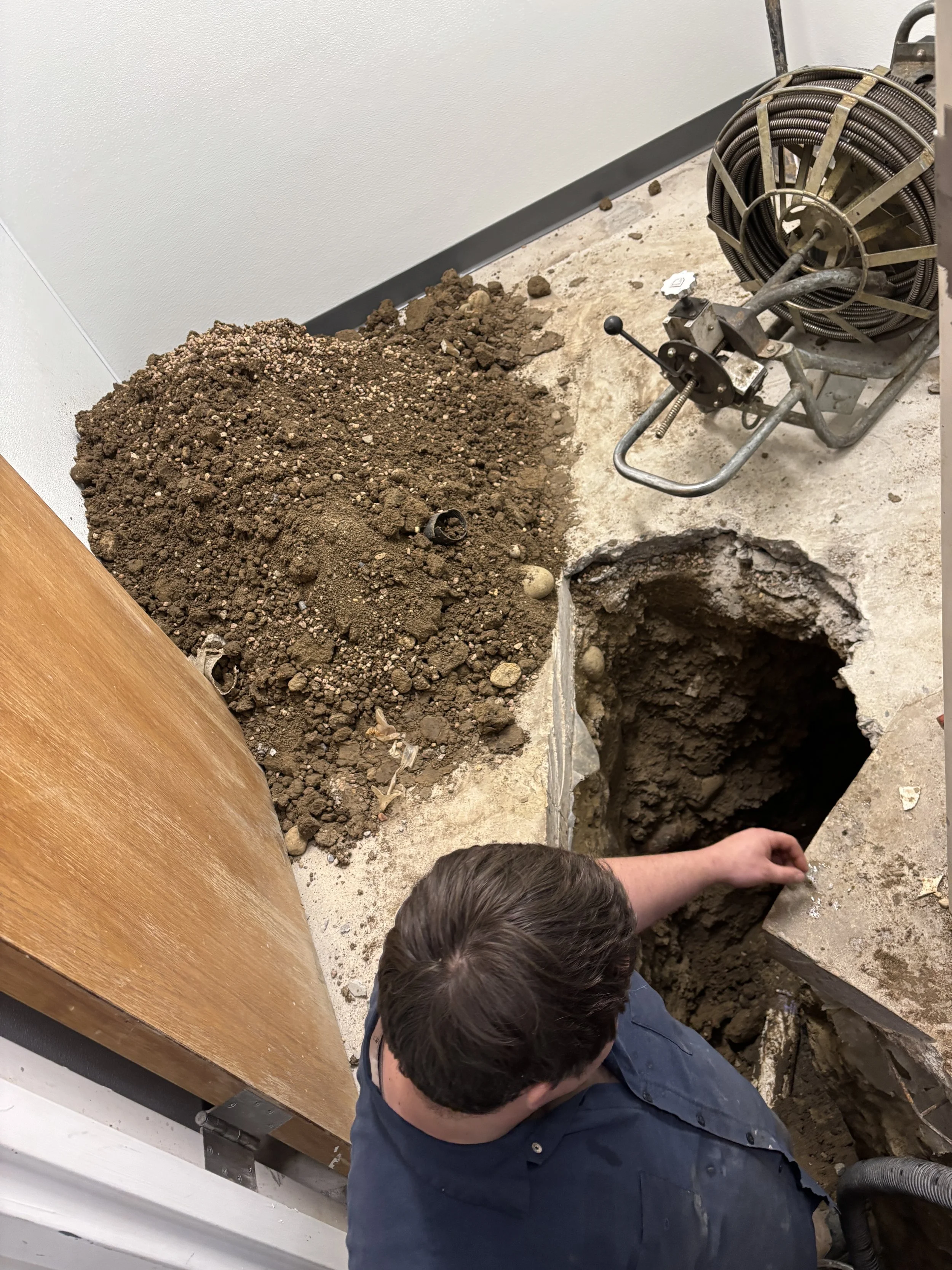 Man digging a hole in the floor of a room, with a pile of dirt next to the hole and a sewer machine nearby.
