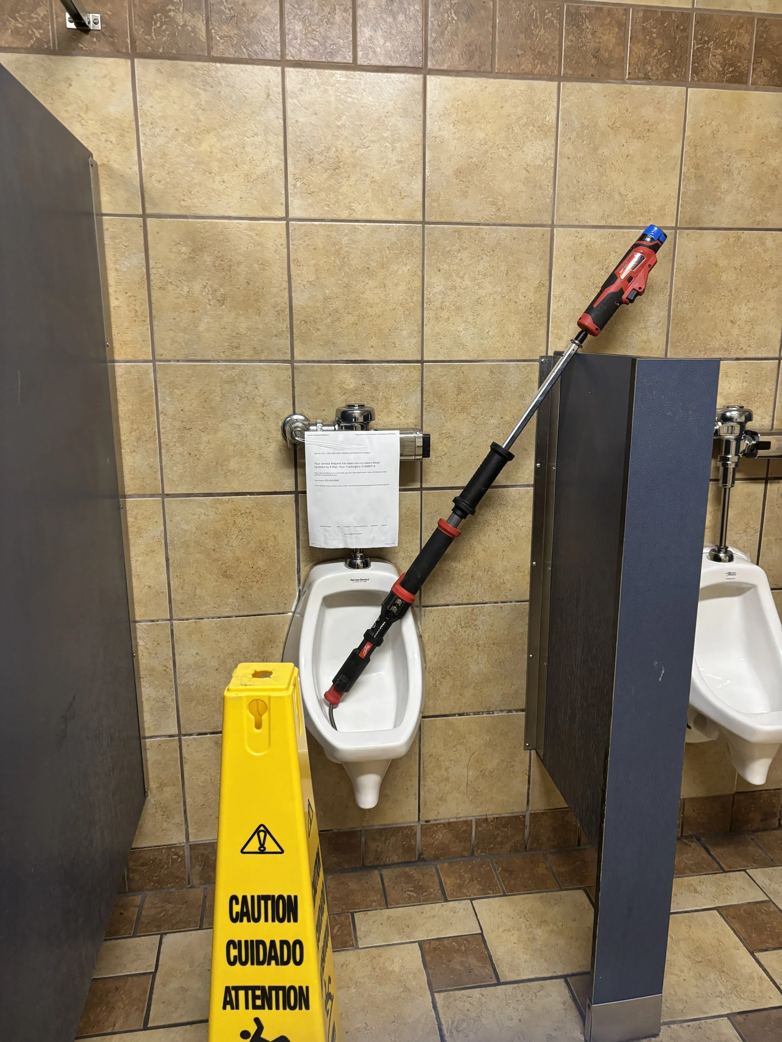 A urinal auger, leaning against a urinal in a public restroom. There is a yellow caution sign on the floor warning about wet floors and a paper attached to the flush pipe above the urinal.
