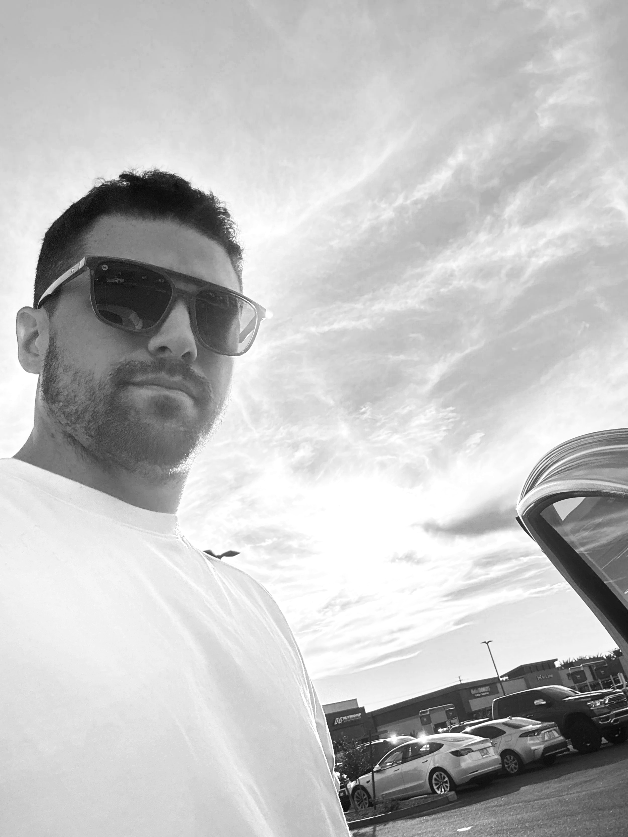 Black and white photo of a man wearing sunglasses and a plain t-shirt, standing outdoors in a parking lot with several cars and a shopping center in the background, under a partly cloudy sky.