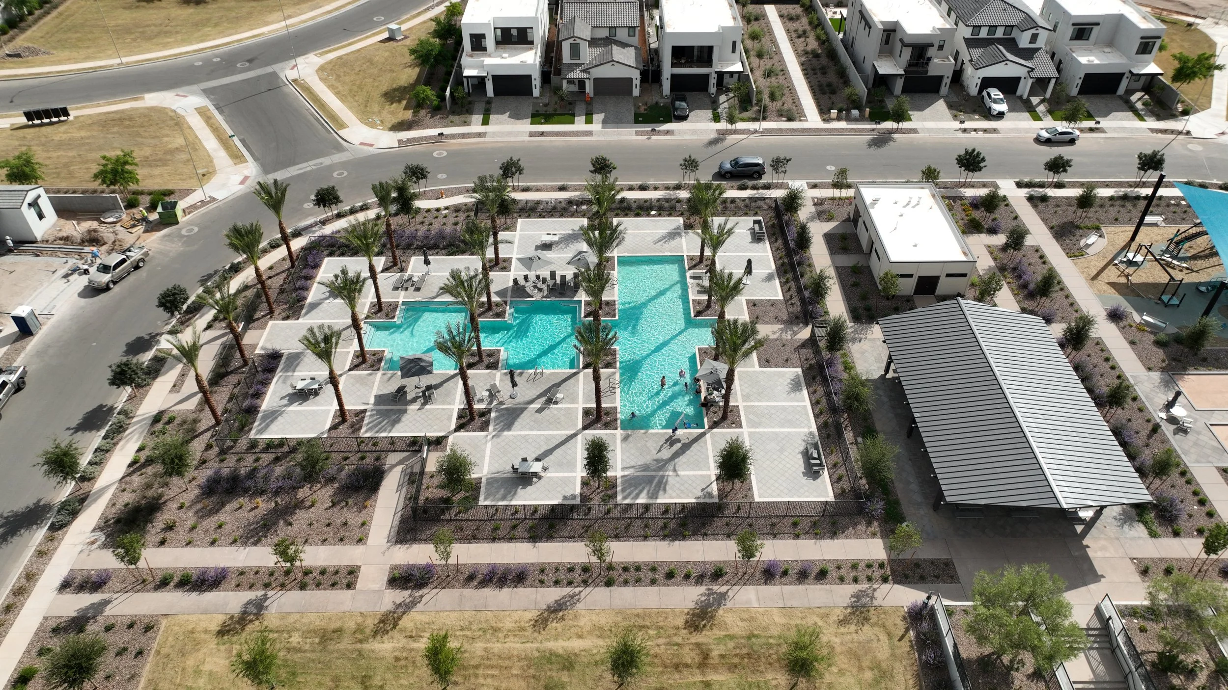 Aerial view of a modern residential community with a central swimming pool surrounded by palm trees, lounge chairs, and pathways. There are houses and landscaped areas around the pool, with a playground and a parking lot nearby.