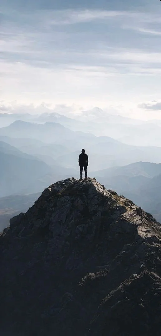 Person standing on a mountain peak looking out over a landscape of mountains and clouds.