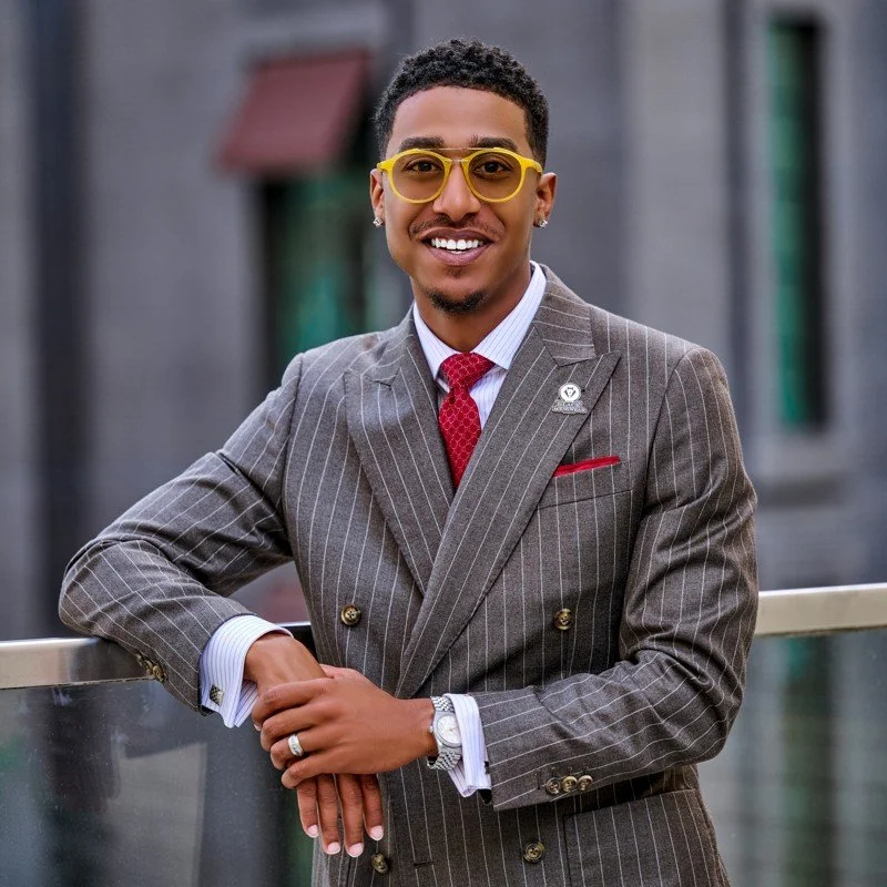 A man dressed in a gray pinstripe suit with a red tie and white shirt, wearing yellow glasses, smiling and leaning on a railing.