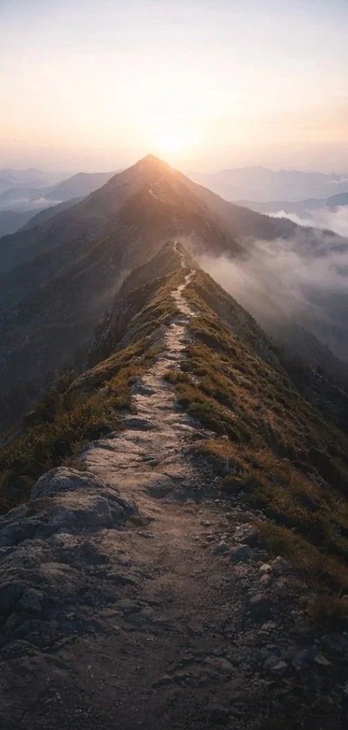 Mountain ridge with a narrow trail leading to a distant peak, overlooking clouds at sunrise or sunset.