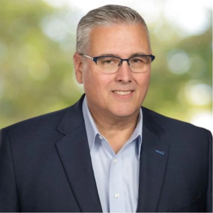 A middle-aged man with gray hair wearing glasses, a light blue shirt, and a dark blazer, standing outdoors with a blurred green background.