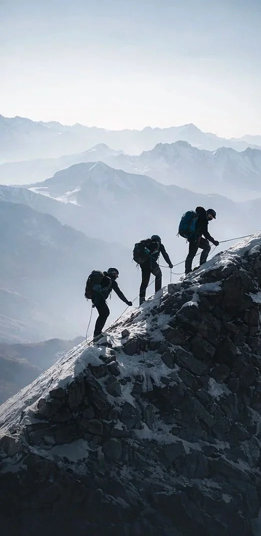 Three mountaineers climbing a snow-covered rocky slope with mountains in the background.