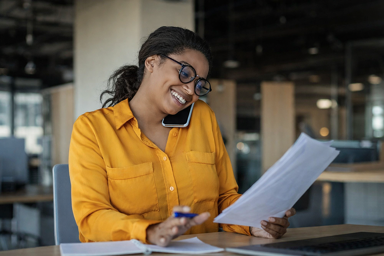 Woman in glasses and yellow shirt smiling while looking at papers in an office.