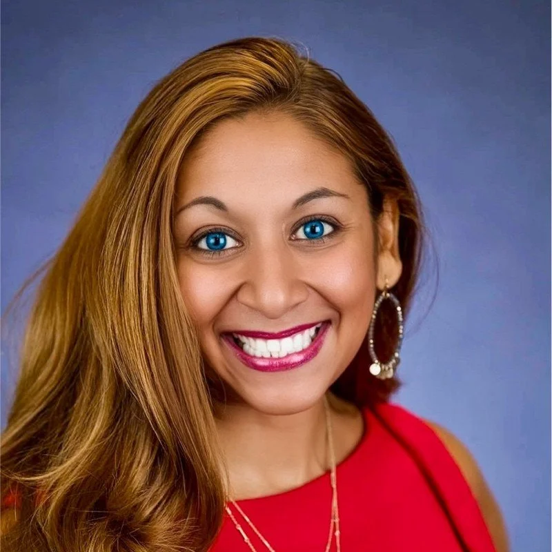 Close-up portrait of a woman with long, wavy, light brown hair, bright blue eyes, wearing a red top, hoop earrings, and a gold necklace, smiling against a blue background.