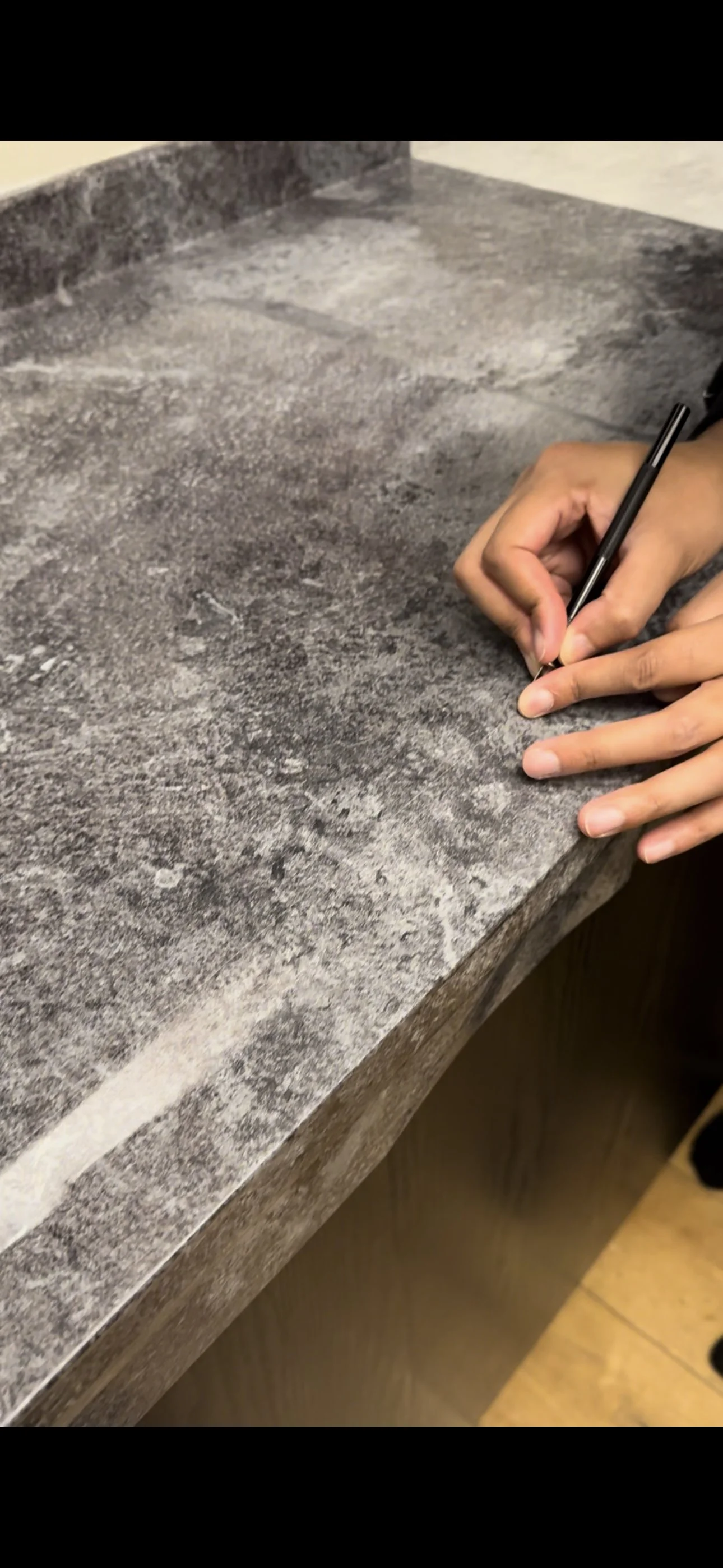 Close-up of a person's hand writing with a pen on a gray, textured stone or concrete surface, possibly a countertop or table.