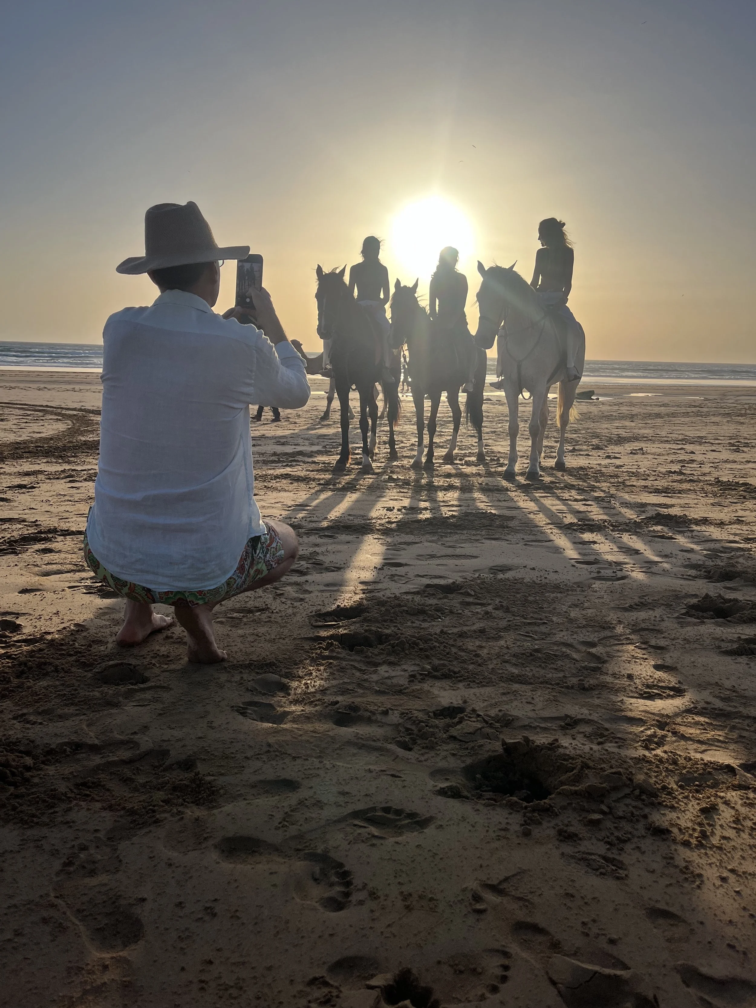 Person in a white shirt, patterned shorts, and a wide-brimmed hat kneeling on the beach taking a photo of four women riding horses at sunset.