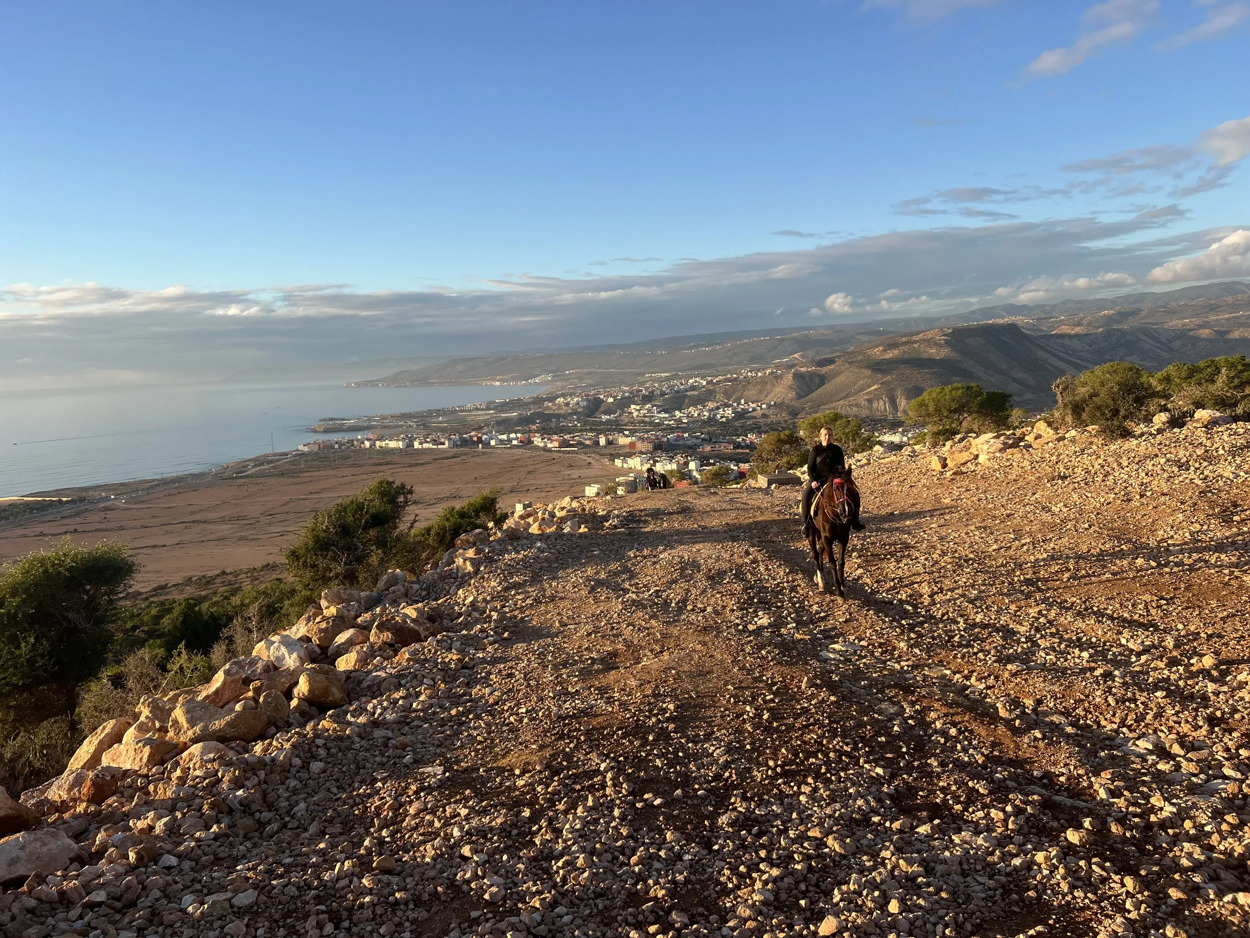 Person riding a horse along a rocky trail on a hillside overlooking a coastal town and ocean at sunset.