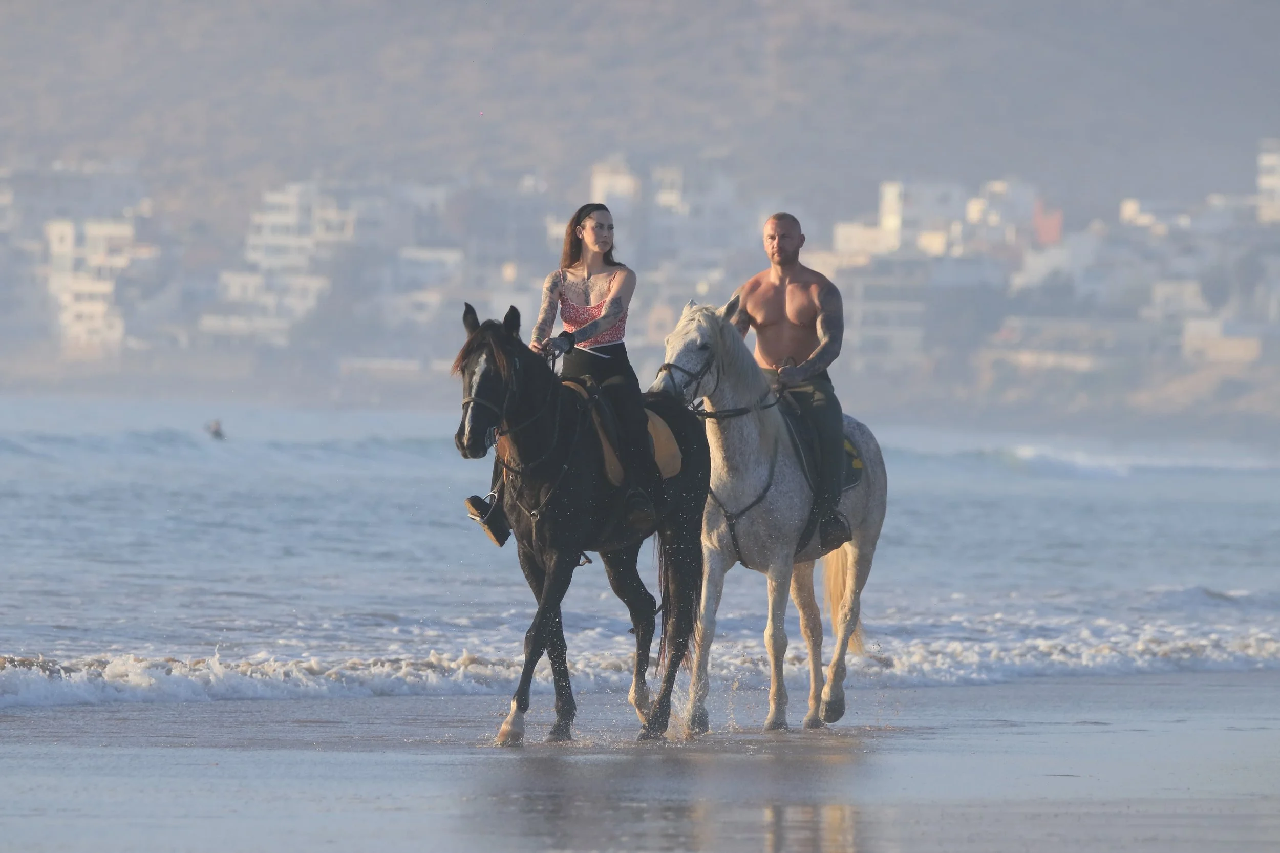 A man and woman riding horses along the beach with ocean waves and city buildings in the background.