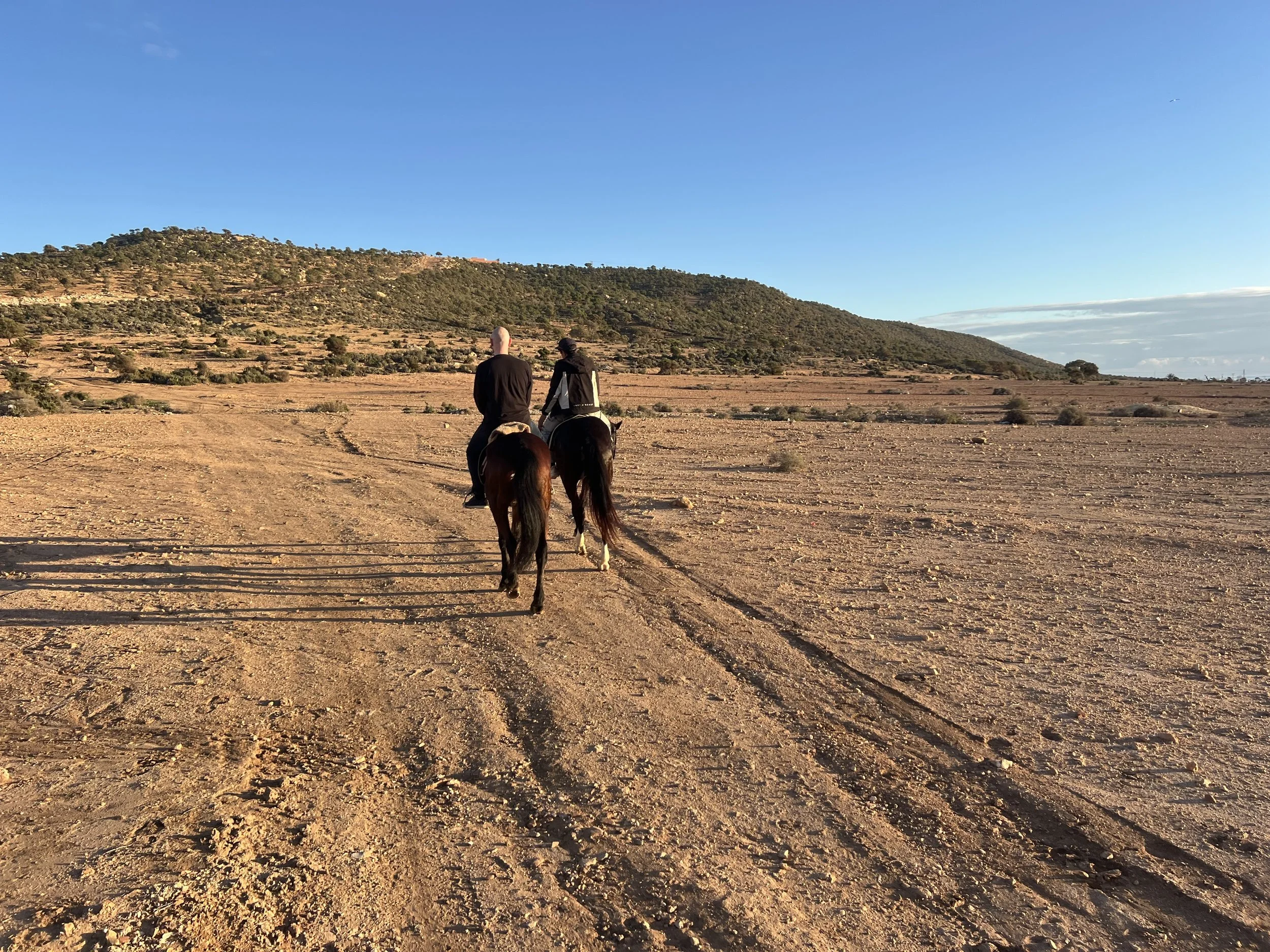 Two people riding horses on a dirt path in a dry, open landscape with hills in the background under a clear blue sky.