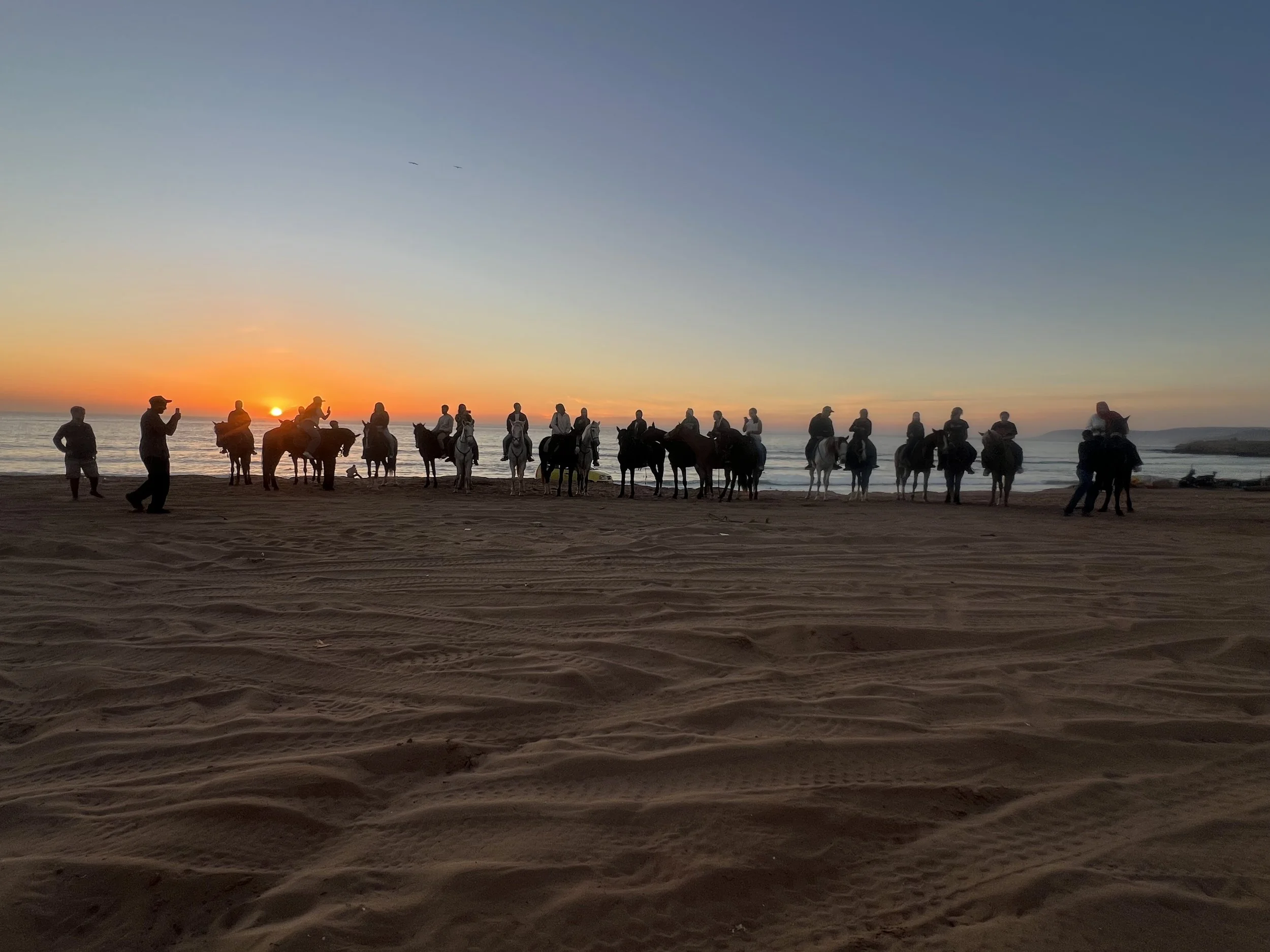 Silhouettes of people riding horses on a beach at sunset with a few individuals standing and taking photos, ocean waves in the background, and a colorful sunset sky.