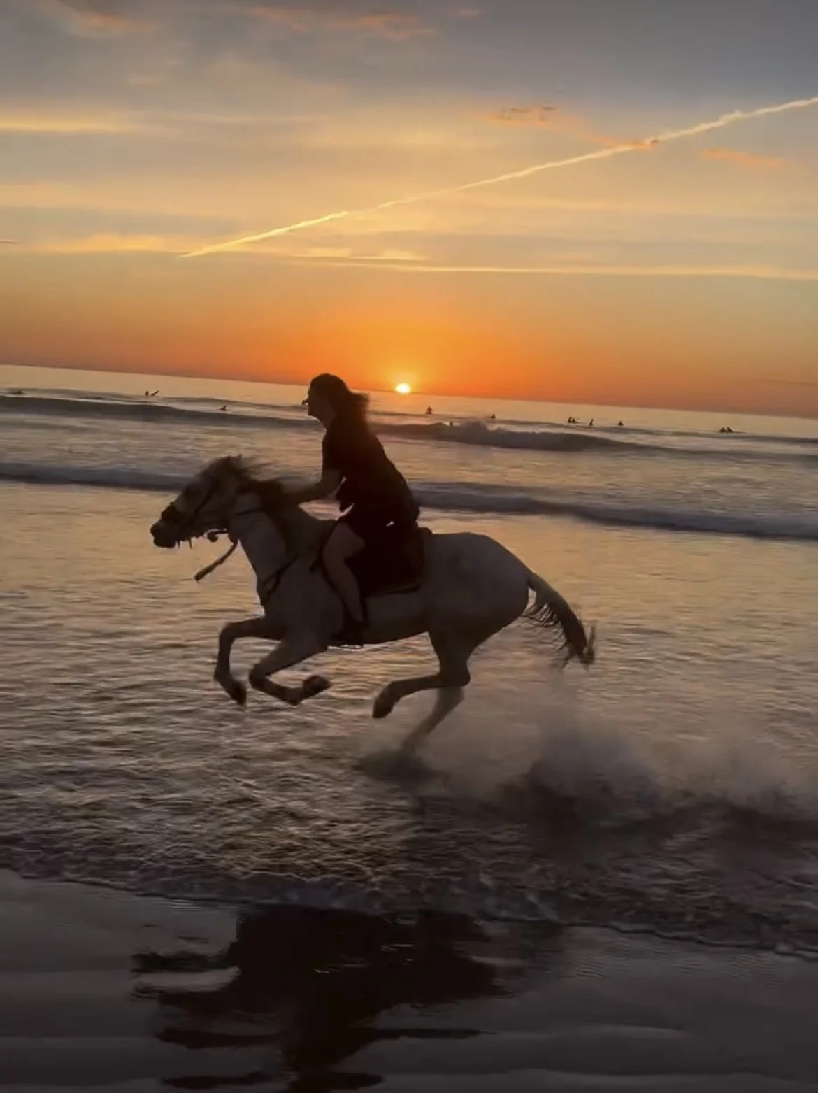 Person riding a horse along the beach at sunset with the ocean and surfers in the background.