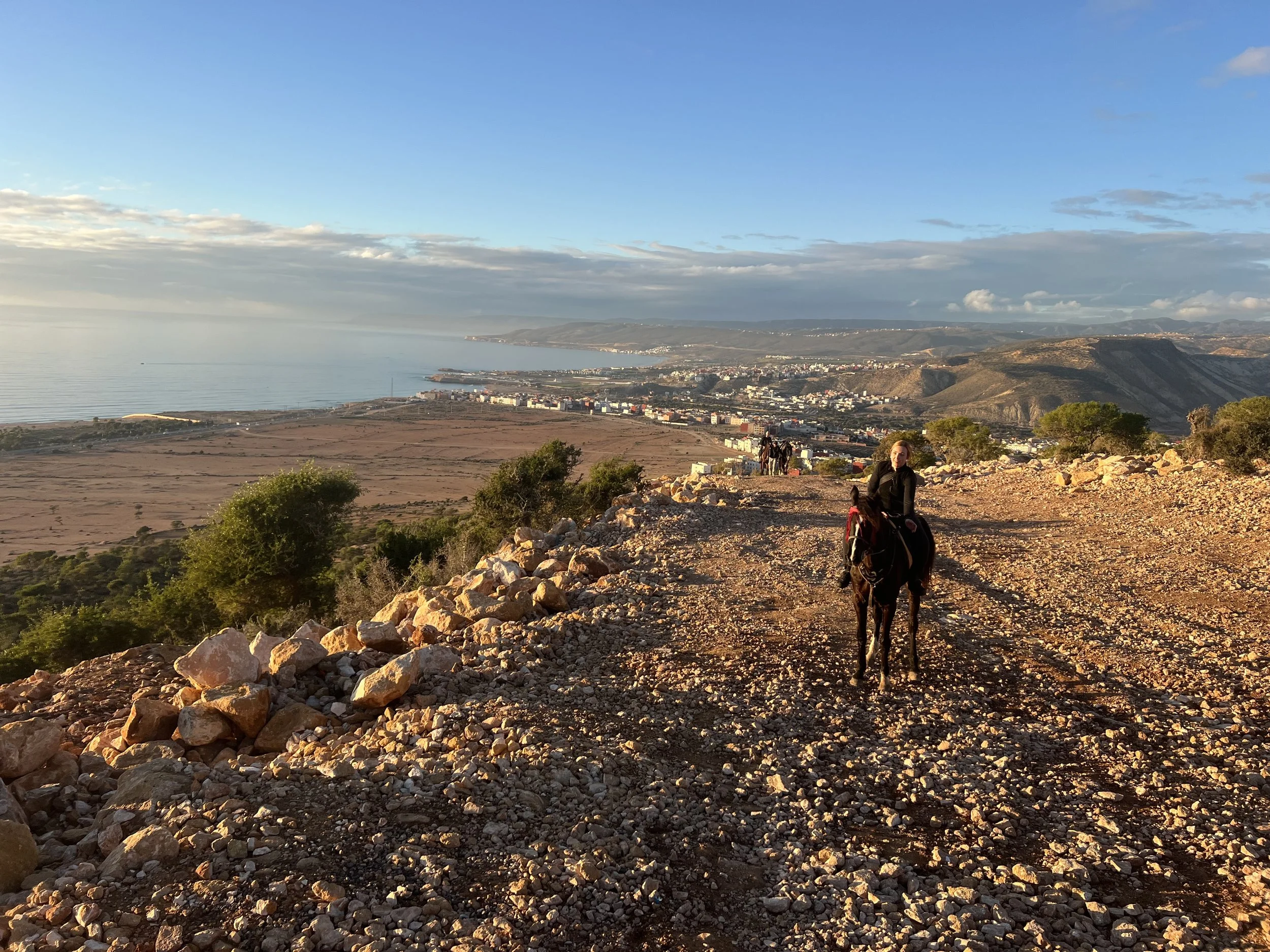 A woman riding a horse on a rocky dirt trail overlooking a coastal town with buildings, hills, and the ocean in the distance during late afternoon or early evening.