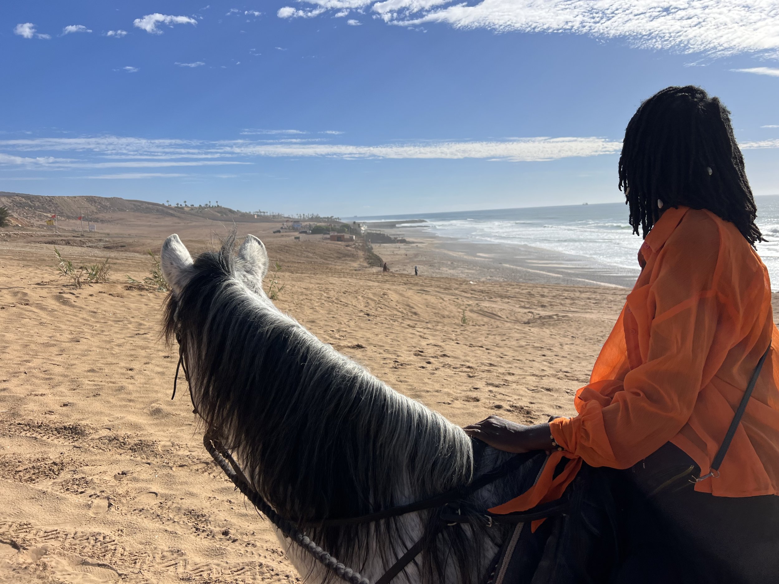 A person with dreadlocks wearing an orange jacket riding a white and black horse on a sandy beach with ocean waves and blue sky in the background.