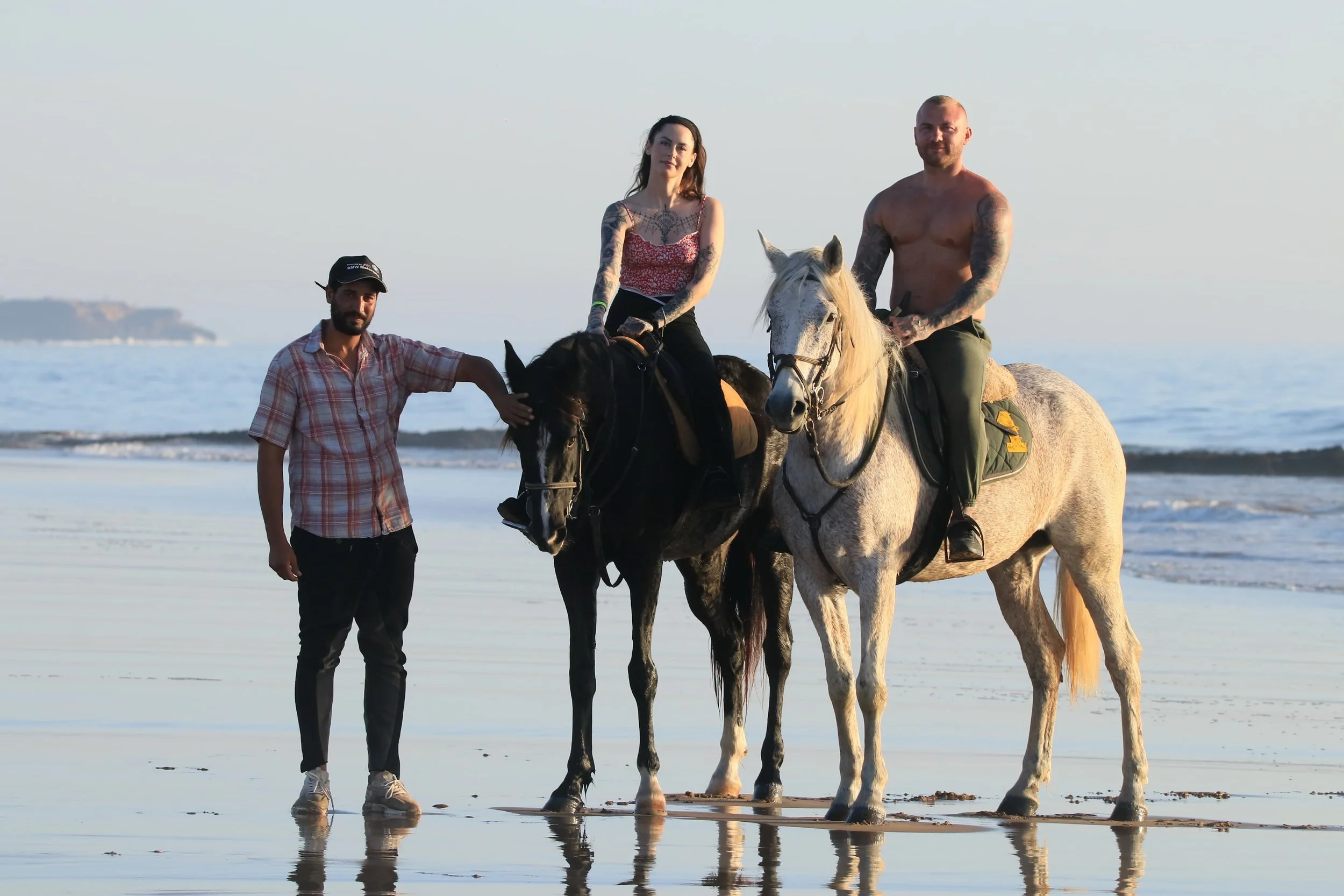 Two men and one woman on horseback at the beach, with a person standing beside one horse, during sunset or sunrise.