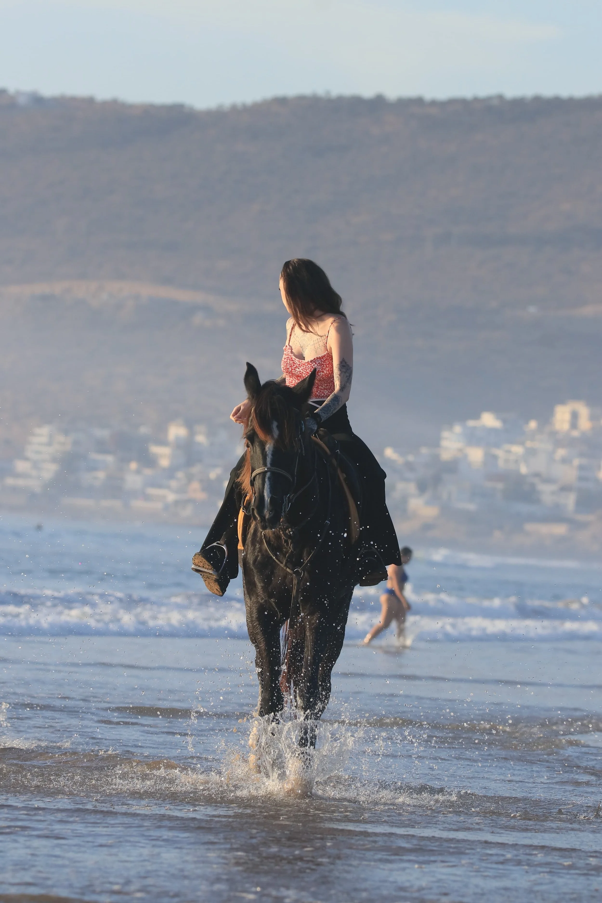 Woman riding a horse in the ocean with people in the background and mountains in the distance.