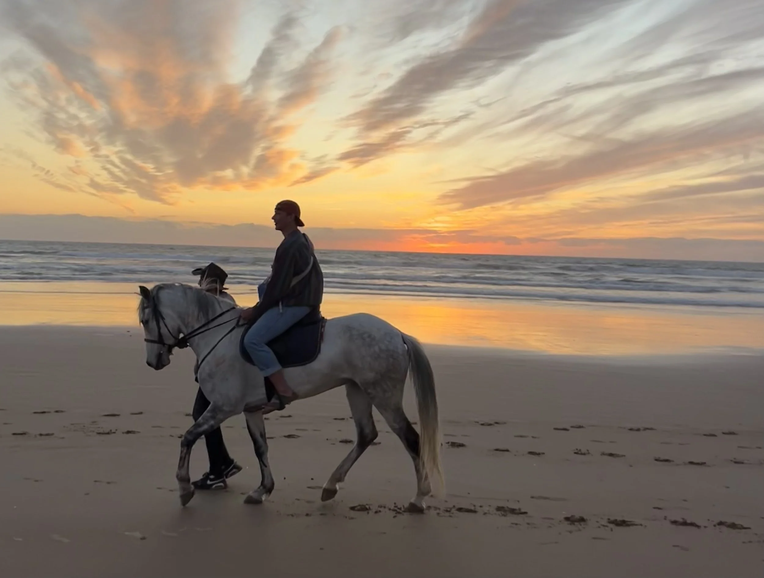 A person riding a white horse along the beach during a colorful sunset with clouds in the sky.