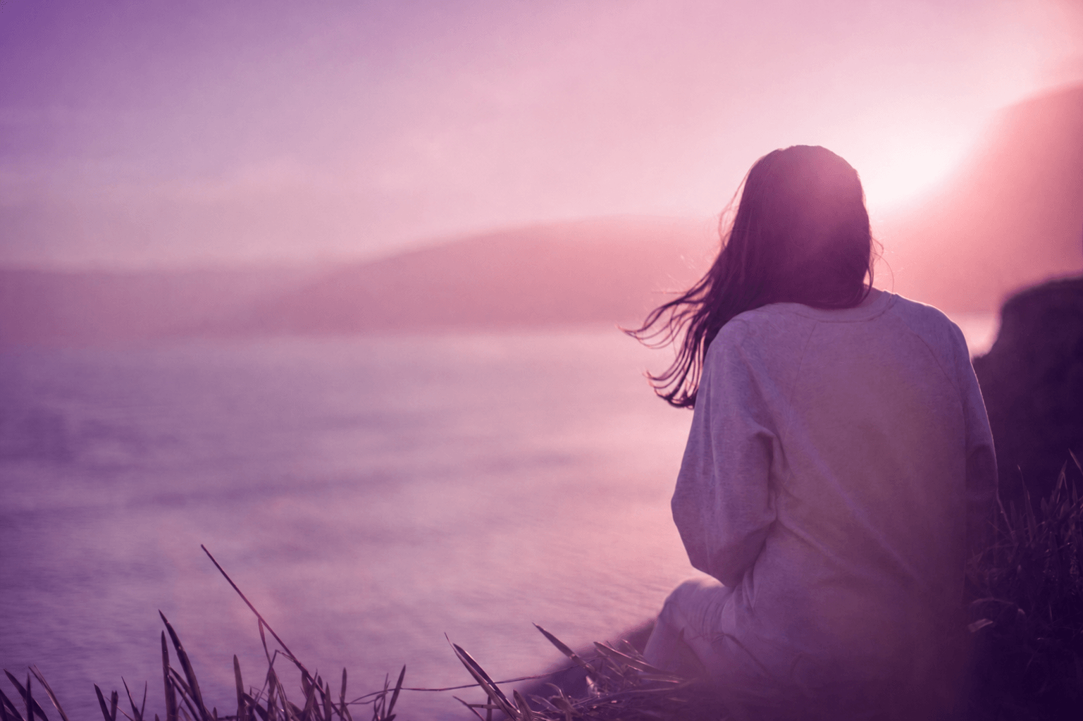 A person with long hair sitting near a body of water during sunset or sunrise, facing away from the camera, with hills or mountains in the background.