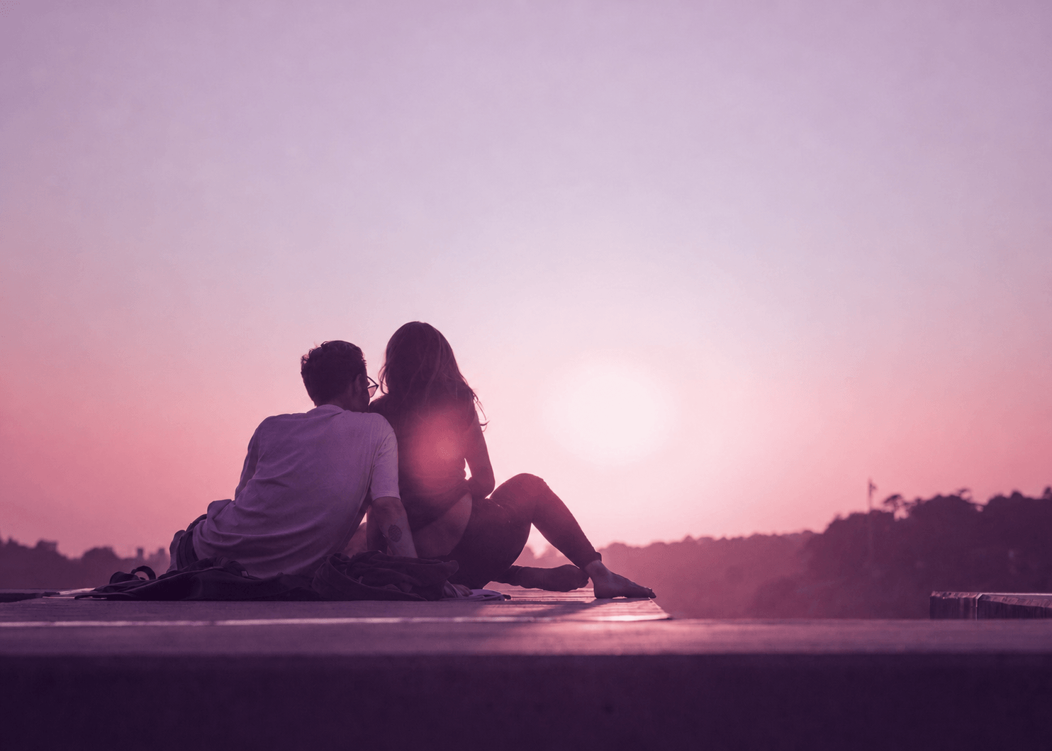 Silhouettes of a young man and woman sitting close together on a dock at sunset, facing the horizon with pink and purple sky.