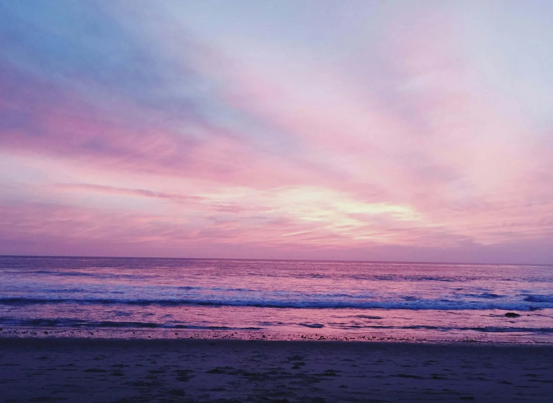 Colorful sunset over the ocean with pink, purple, and blue hues in the sky and gentle waves at the shoreline.