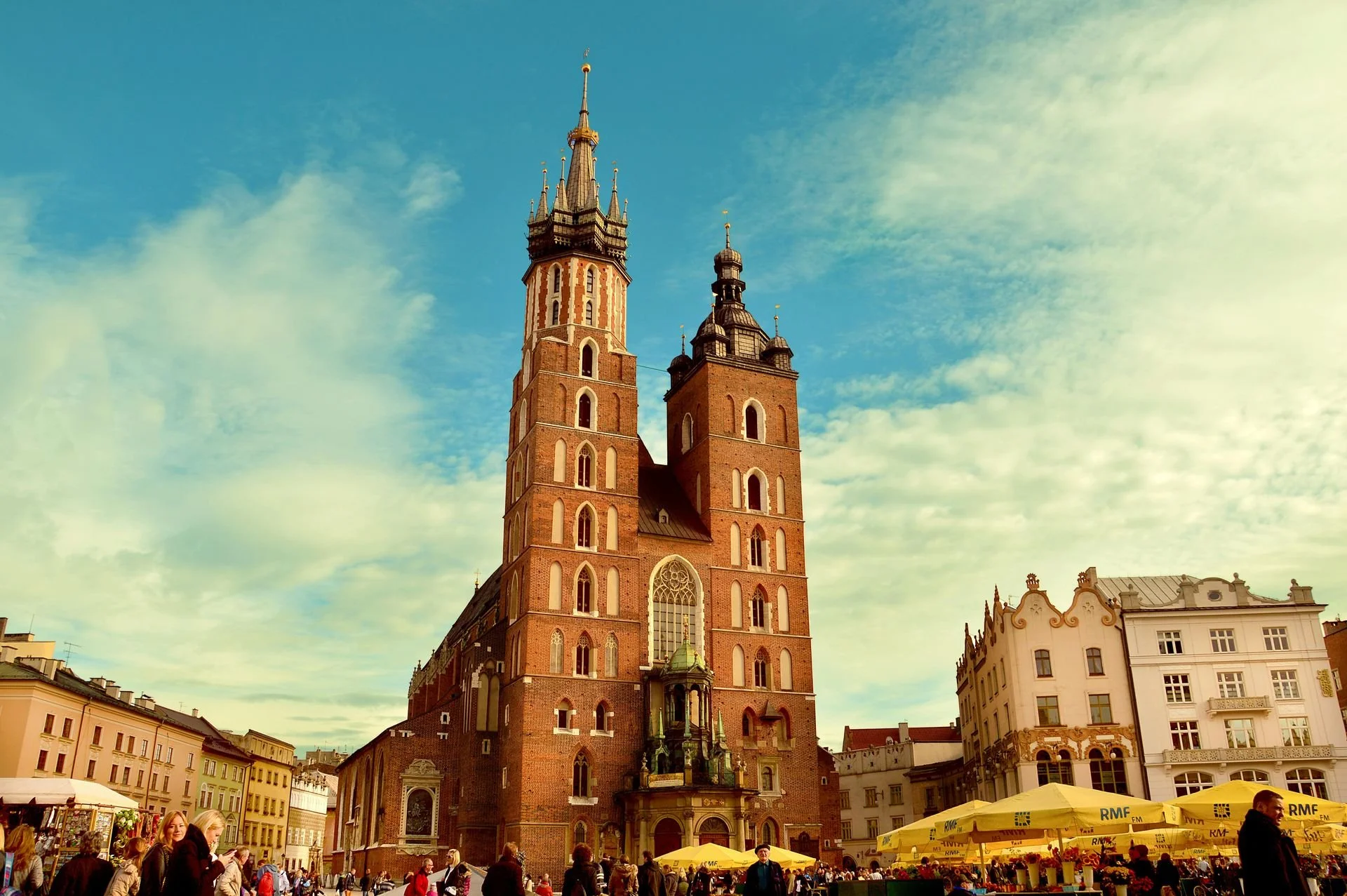 Tall brick church with Gothic architecture, twin spires, and a large arched window, surrounded by a lively market square with yellow umbrellas, people, and historic buildings under a blue sky with scattered clouds.