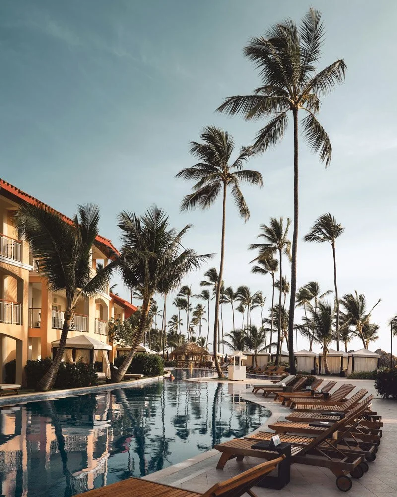 Vacation resort pool area with several lounge chairs, palm trees, and a building with balconies, under a clear sky.