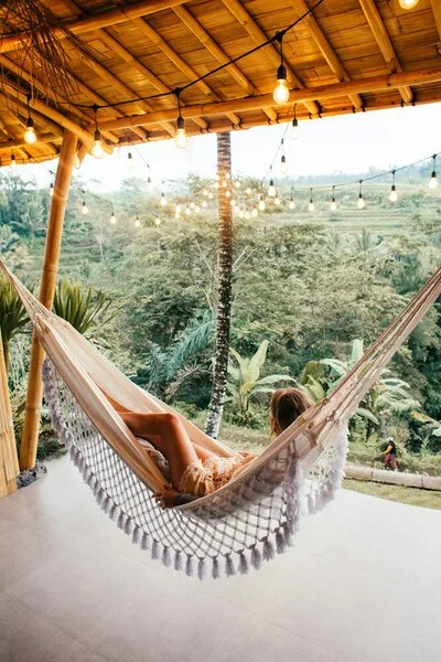 Person relaxing in a hammock on a porch overlooking a lush, green landscape with trees and a person riding a bicycle in the distance, under a wooden roof with hanging string lights.