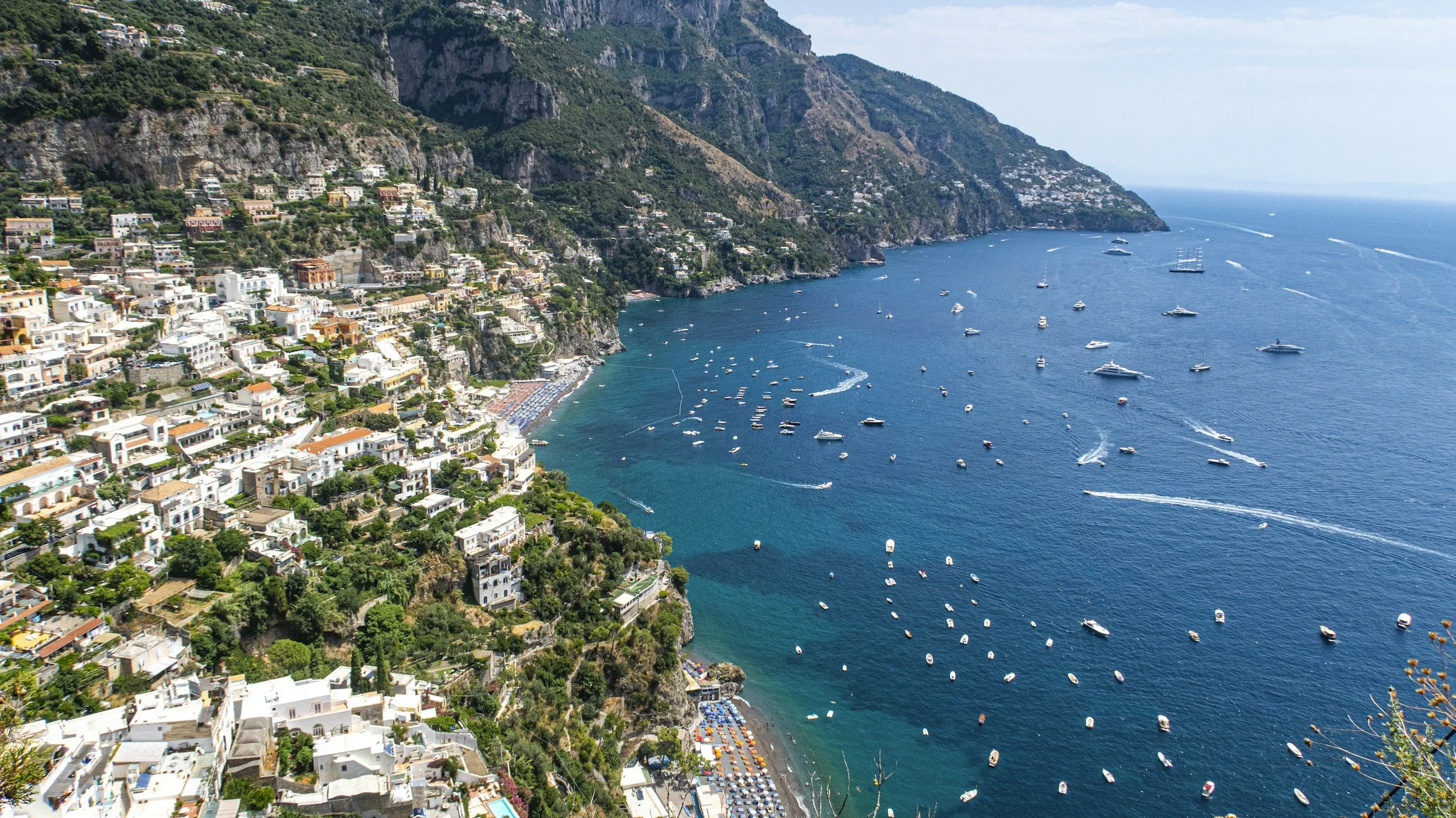 A coastal town on steep cliffs with white and pastel-colored buildings overlooking a blue ocean filled with boats and yachts, with lush green mountains in the background.